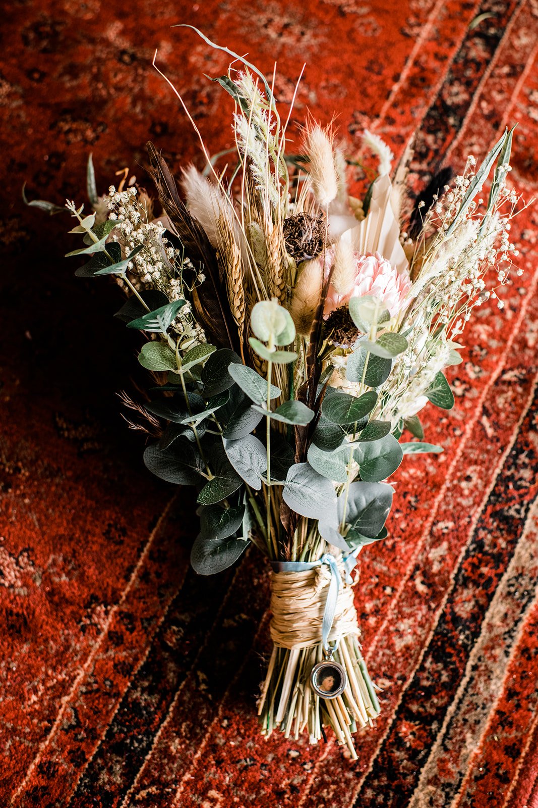 A bridal bouquet of pale flowers, dried grasses and eucalyptus rests on a patterned rug.