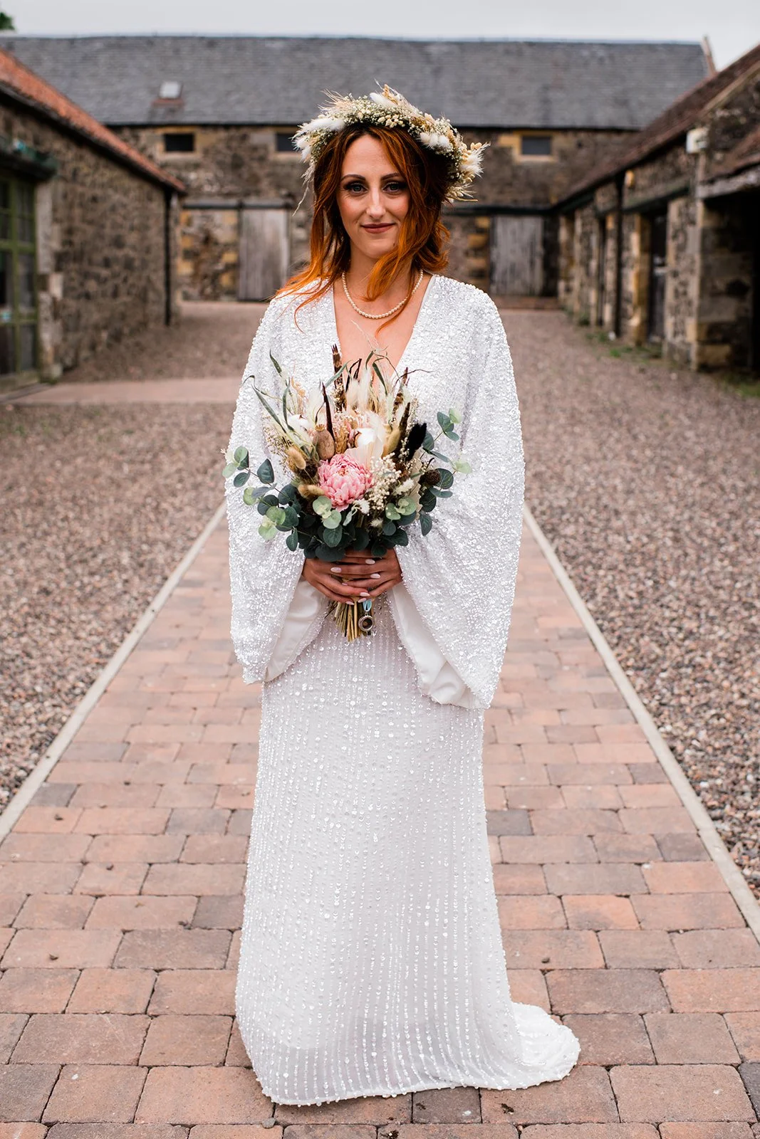 The bride stands in Pratis Barns’ stone courtyard, holding her bouquet and wearing a floral crown.