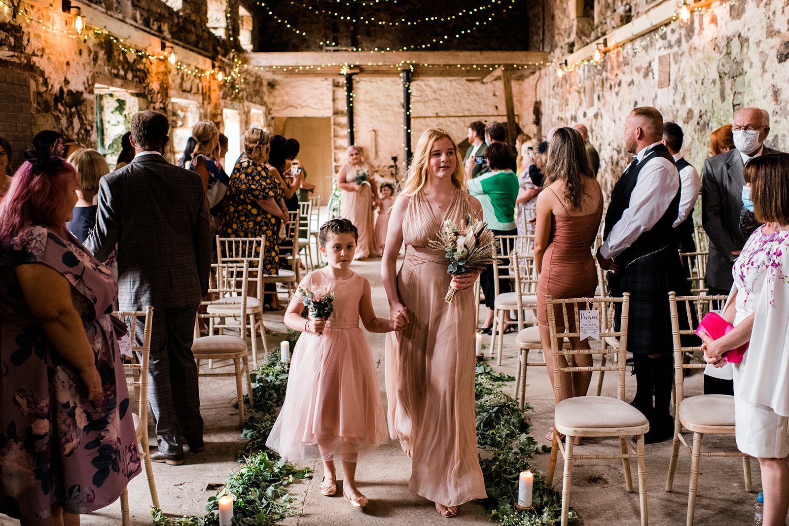 Flower girls walk down the aisle between standing wedding guests inside the barn.