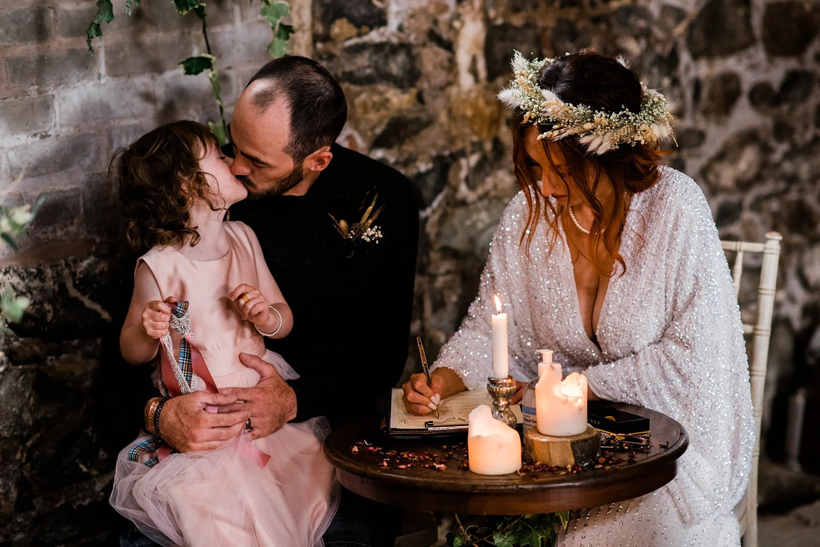 The groom kisses a young flower girl as the bride smiles beside them.