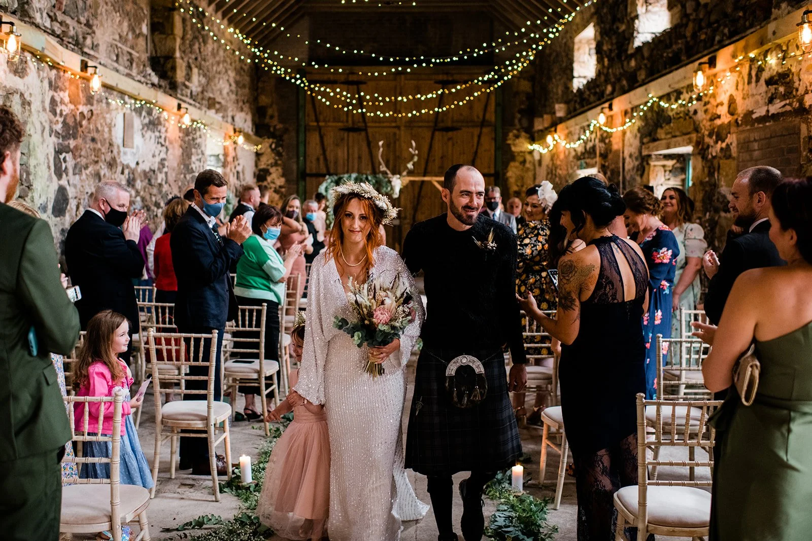 The newly married couple walk down the barn aisle with a flower girl as guests celebrate.