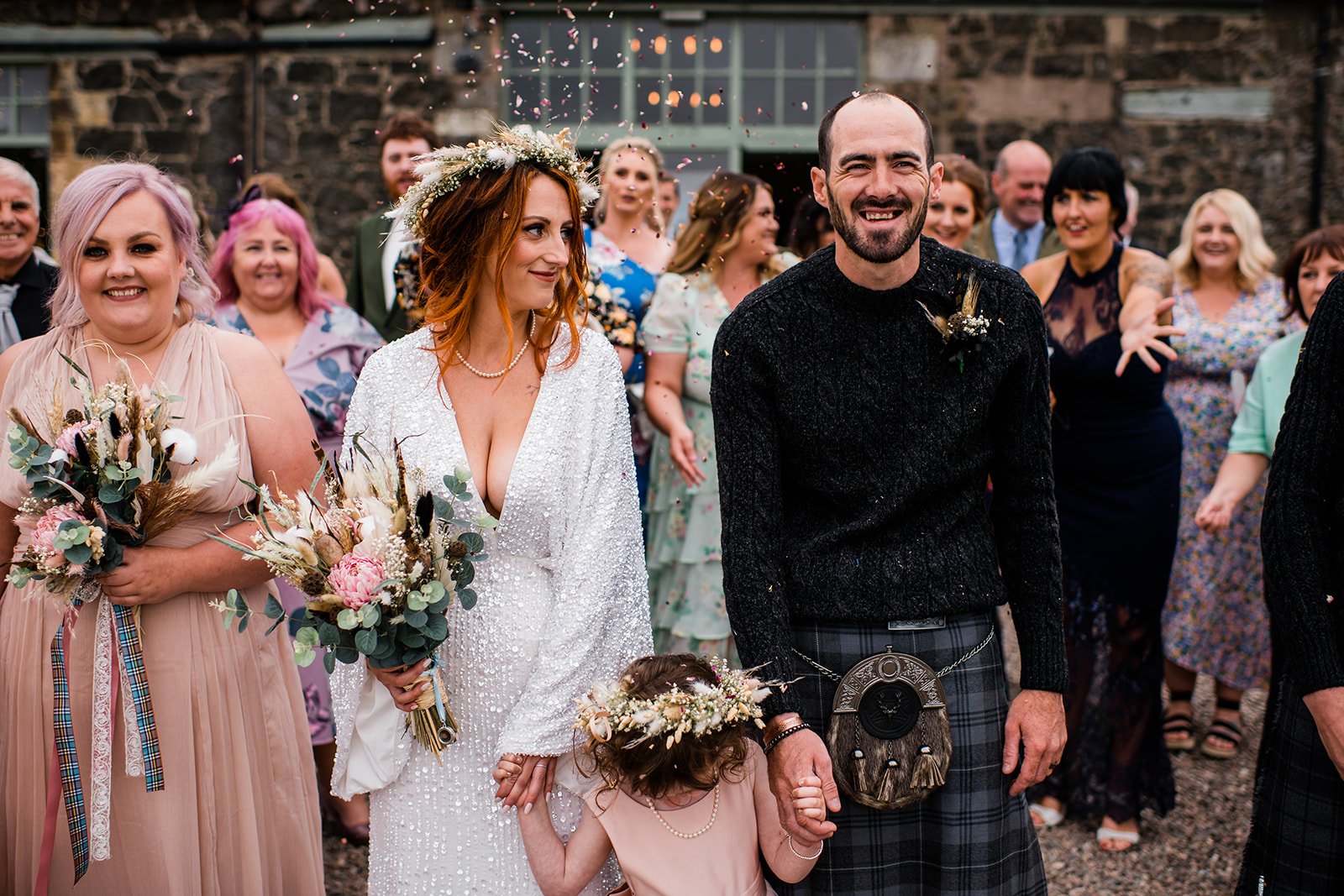The bride and groom hold hands with a young flower girl as guests throw confetti outside Pratis Barns.