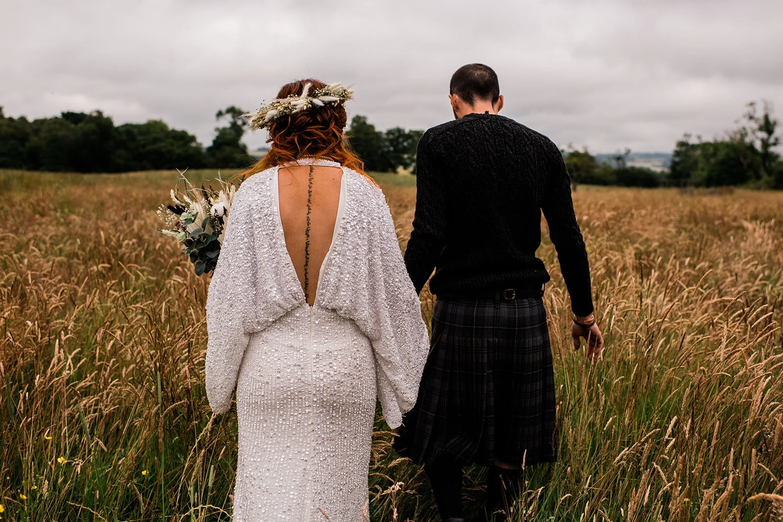 The couple walk hand in hand away from the camera through a meadow.