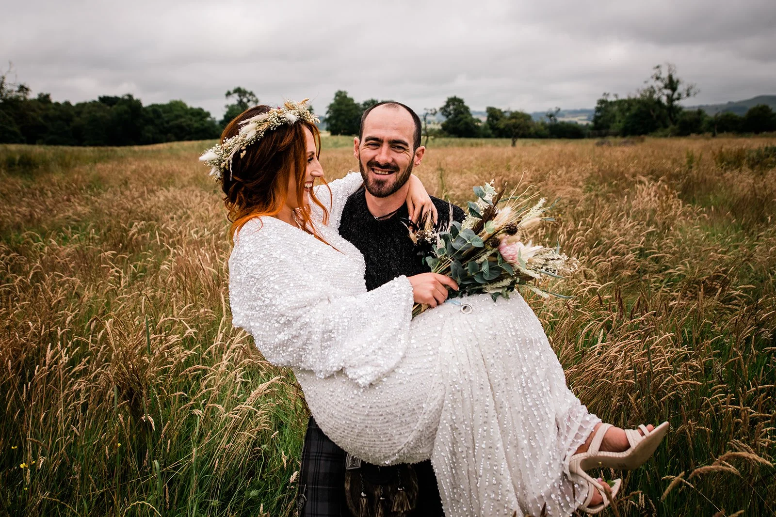 The groom carries the smiling bride through a meadow at Pratis Barns, with her bouquet in her lap.