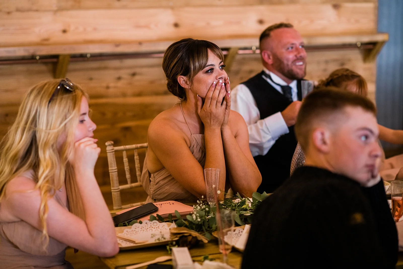 Guests smile and react to a speech at the wedding reception tables.