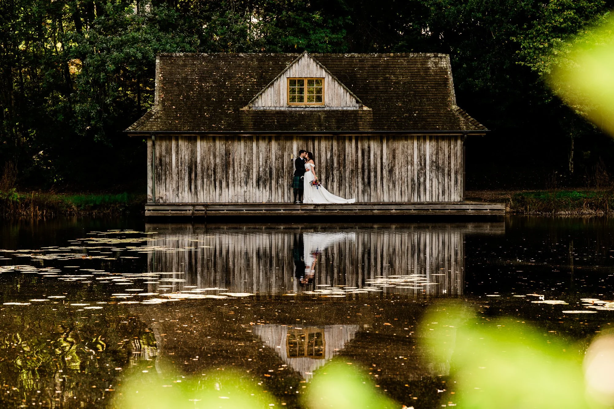 Aimee and Matthew outside the wooden Boathouse at Kinmount House, reflected in the still water.