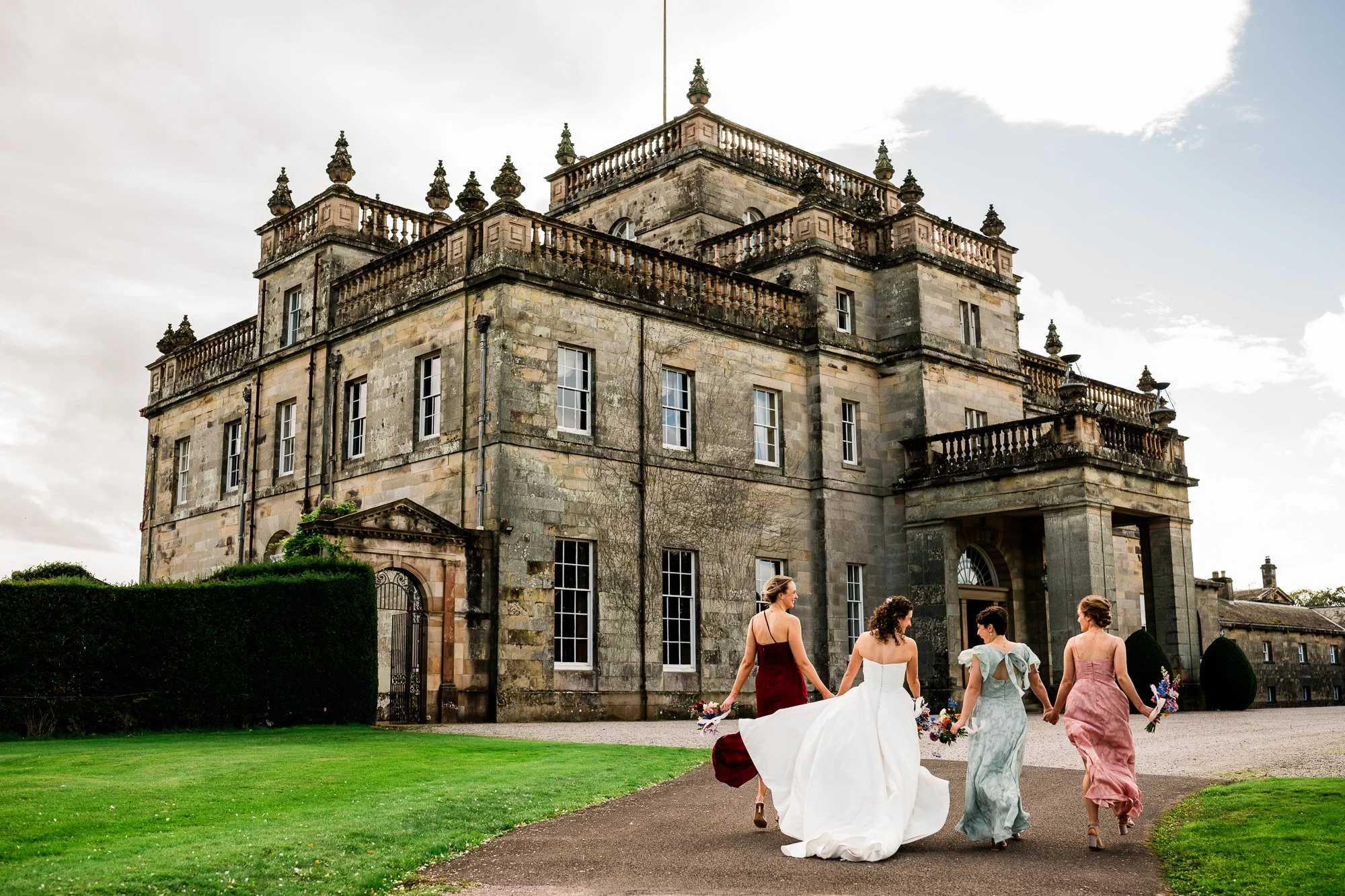 Aimee and her bridesmaids walk towards Kinmount House, carrying bouquets and holding the bride’s train.
