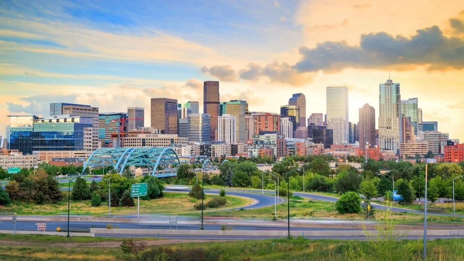 A city skyline with tall buildings during sunset, with a highway and green trees in the foreground.