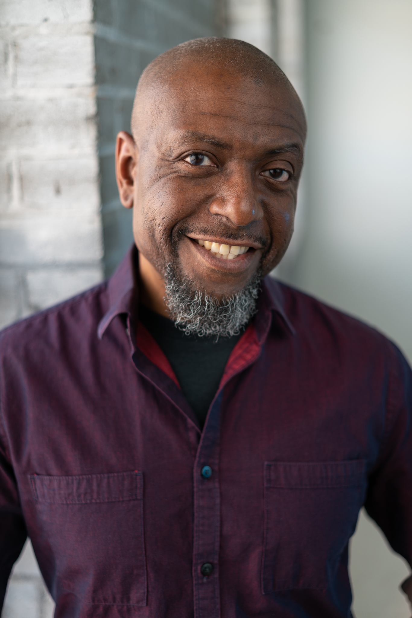 A smiling man with a beard wearing a maroon shirt standing next to a gray brick wall.