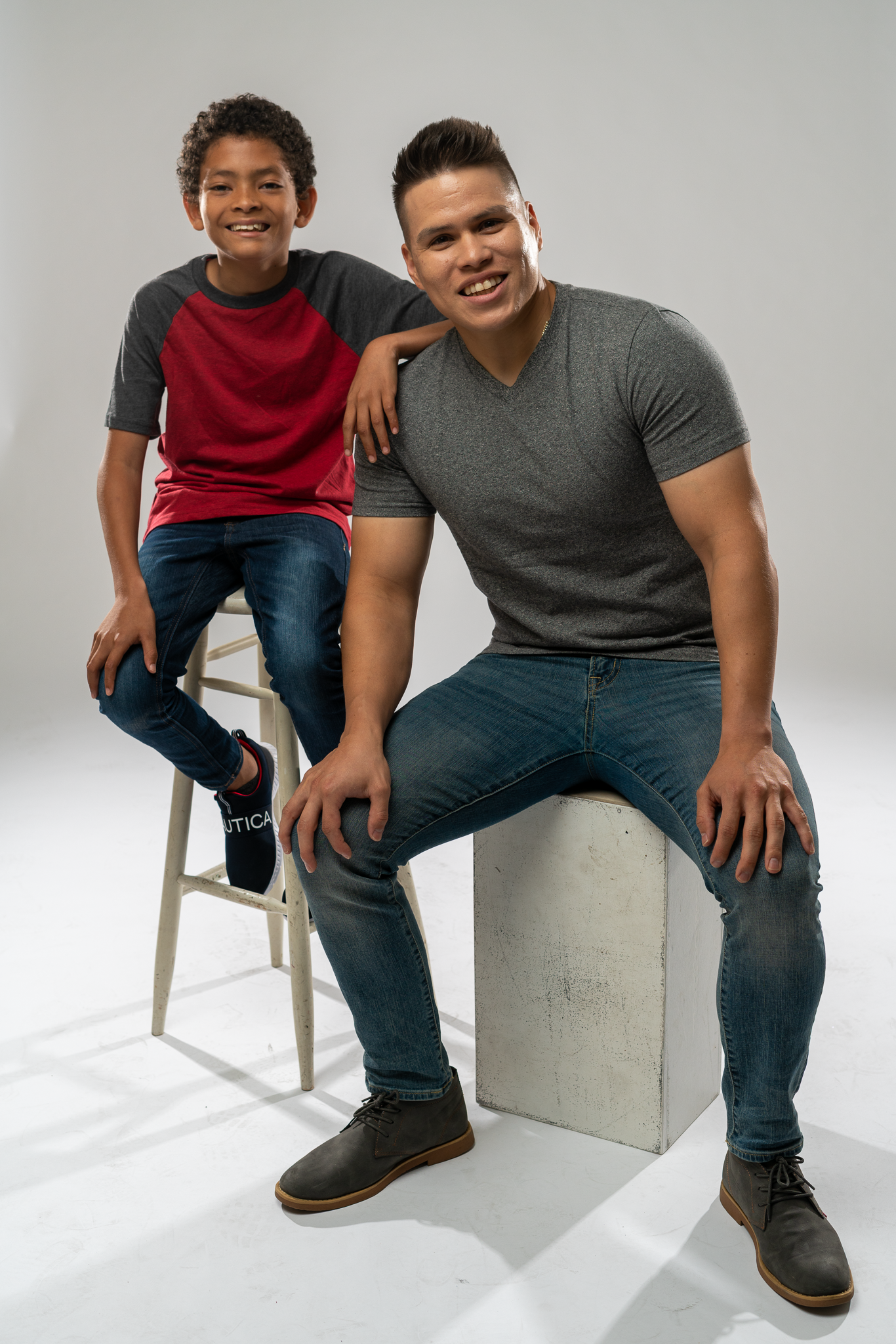 Two smiling males, one child and one adult, posing together in a photo studio with a plain background. The child is sitting on a stool while the adult is sitting on a concrete block, with the adult's arm resting on the child's shoulder.