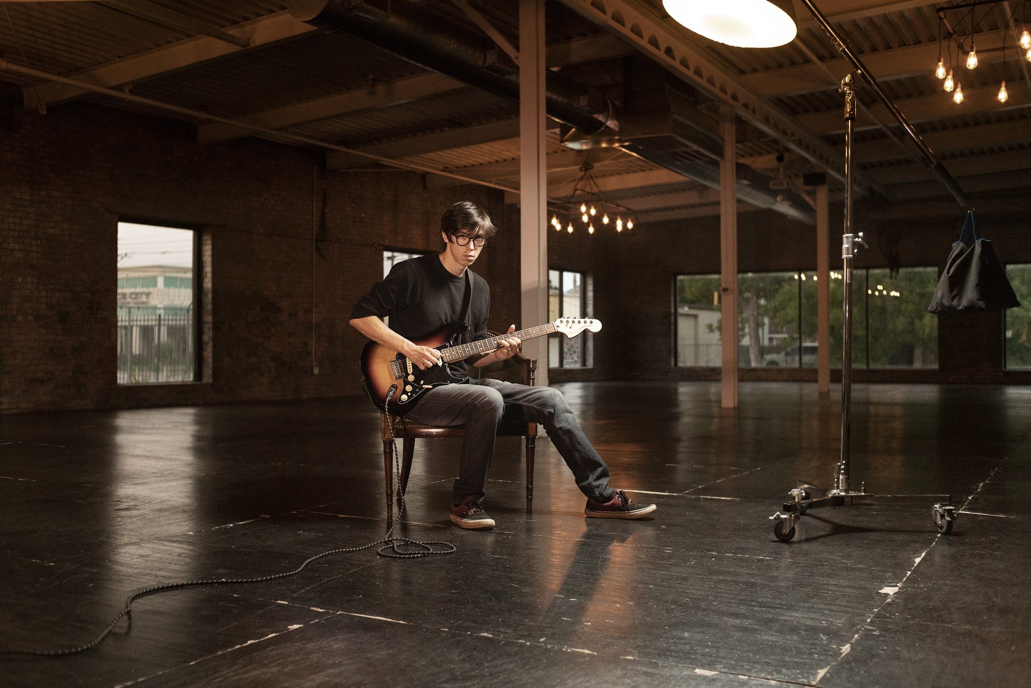 A young man sitting on a chair in an empty industrial studio, playing an electric guitar, with studio equipment and large windows in the background.