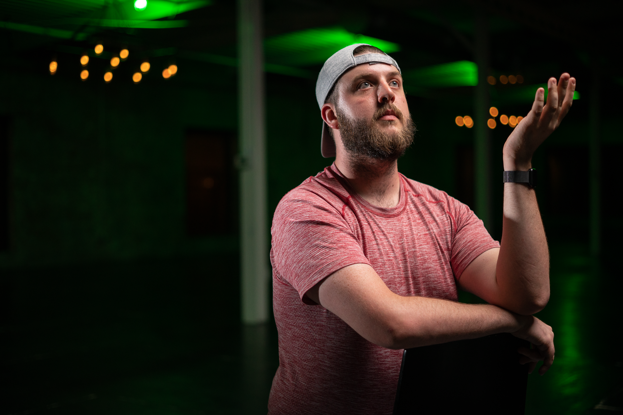 A bearded man wearing a gray cap and red t-shirt stands with one arm crossed and the other raised, in a dimly lit room with green lighting and string lights.