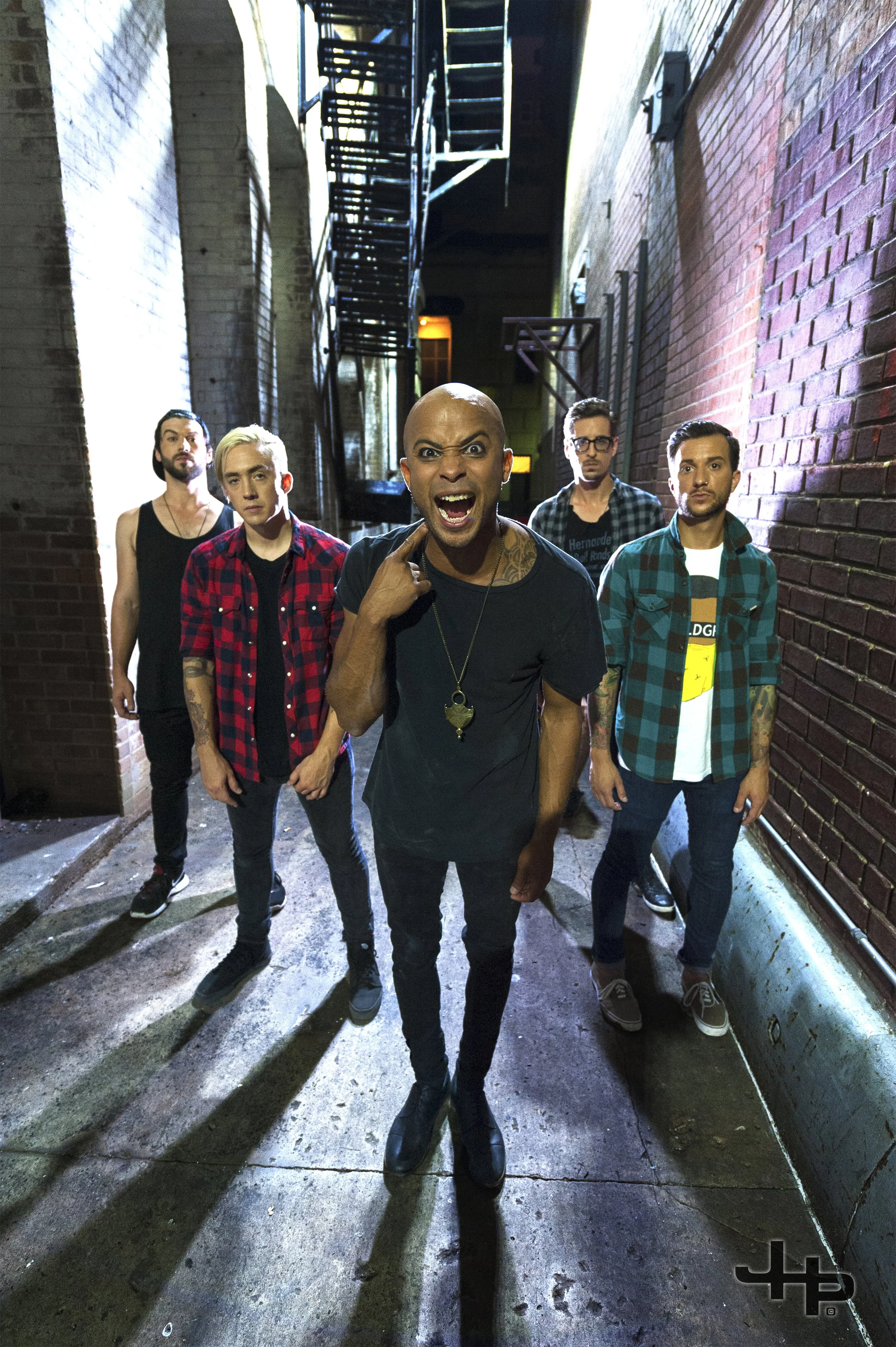 Group of five men standing in a narrow alleyway with brick walls on each side. The man in the front, who is bald and has tattoos, is shouting and pointing to his face. The others are standing behind him, looking at the camera.