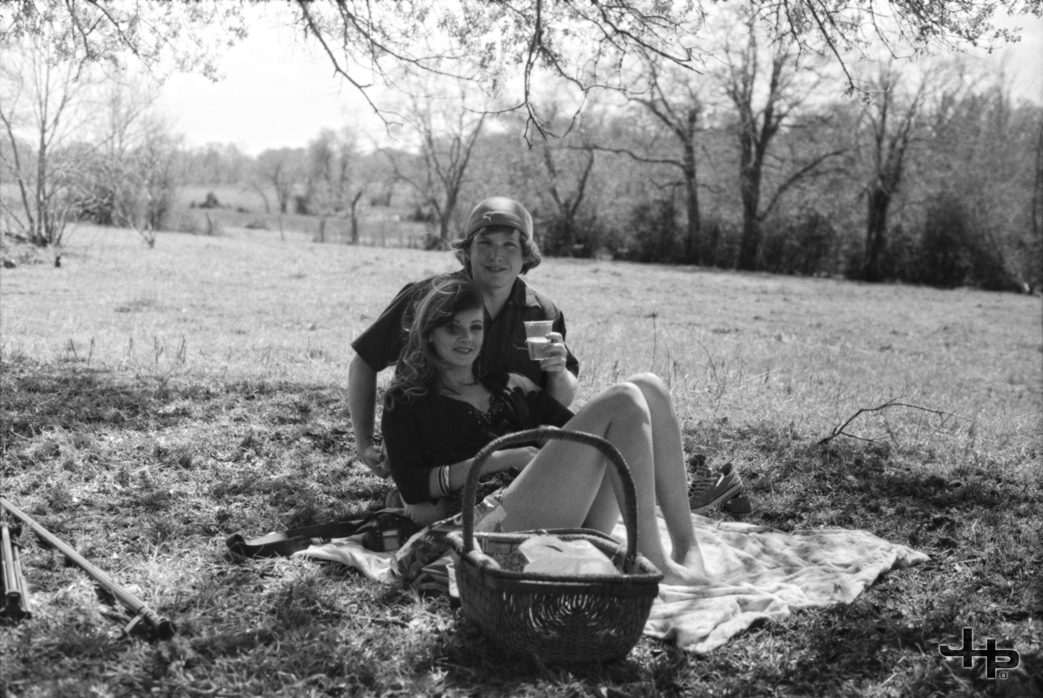 A black and white photo of a young man and woman sitting on a blanket in a grassy field, with the man holding a cup and both smiling. There are trees in the background and items like a basket, a pair of shoes, and a camera nearby.