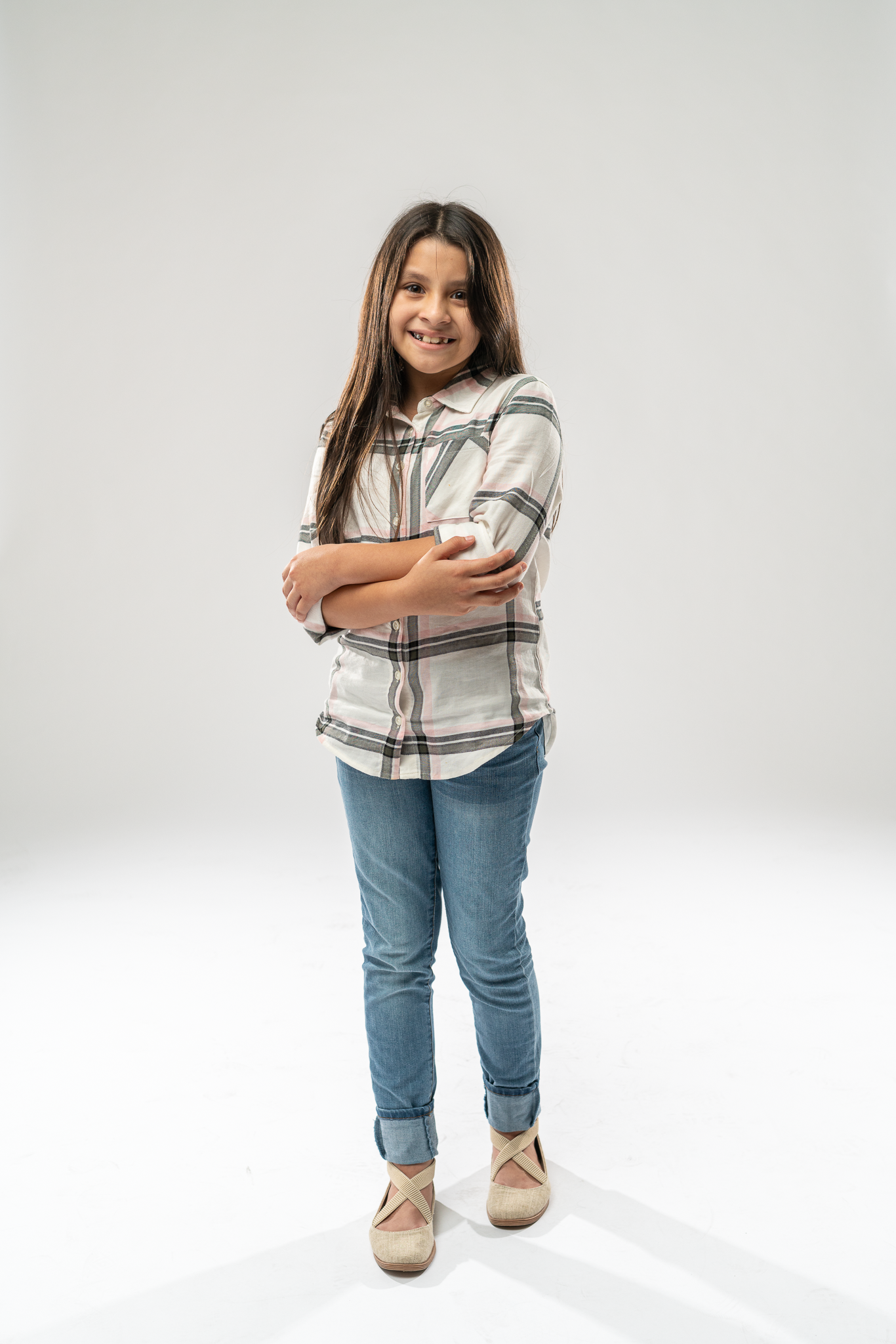 A young girl with long brown hair, wearing a plaid shirt and jeans, smiling with arms crossed, standing against a plain white background.