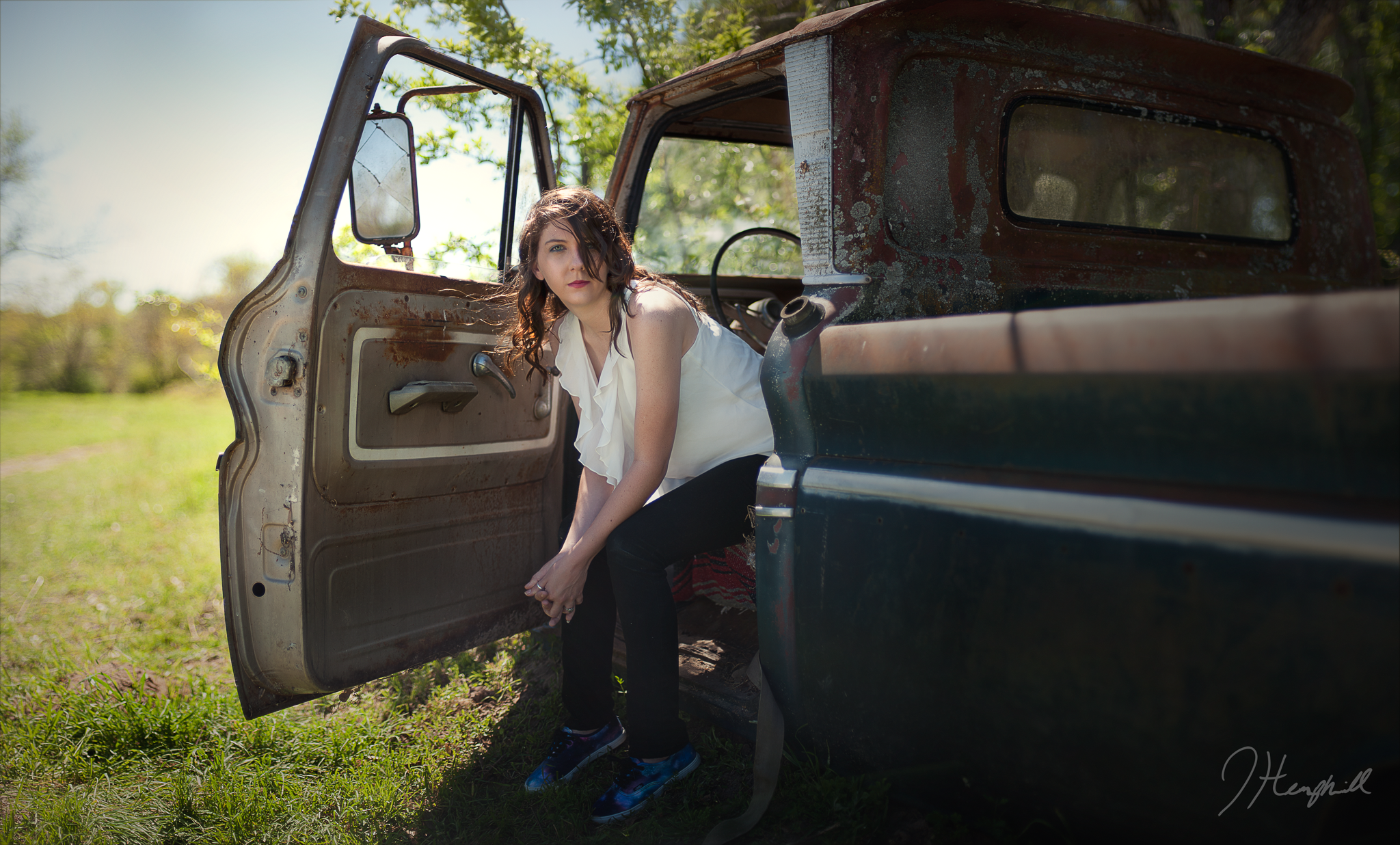 A woman with wavy hair wearing a white sleeveless top leaning into an old rusty pickup truck with open door. She is outdoors on green grass with trees in the background, under a clear sky.