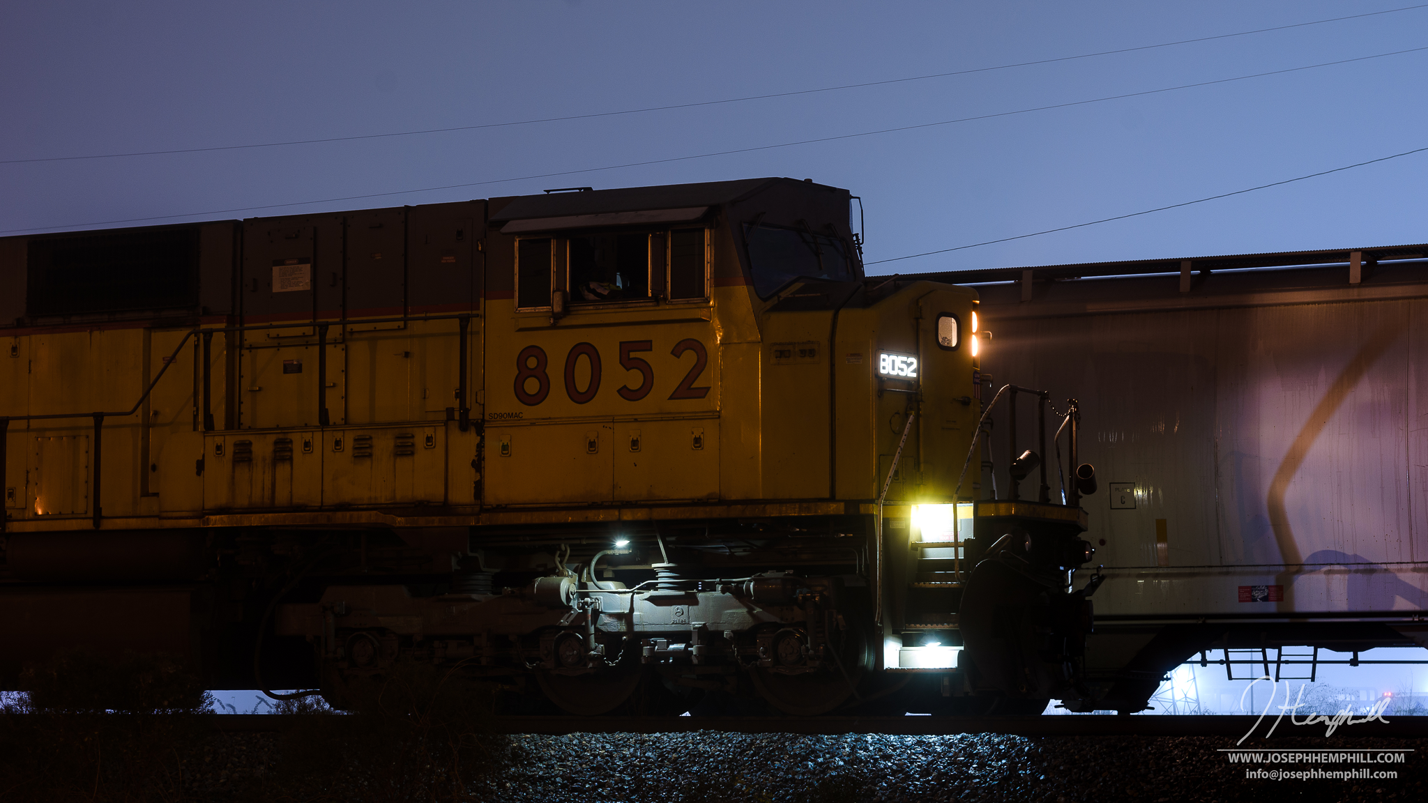 Nighttime photo of a yellow locomotive train with the number 8052, illuminated by headlamps, moving along tracks with a clear sky in the background.