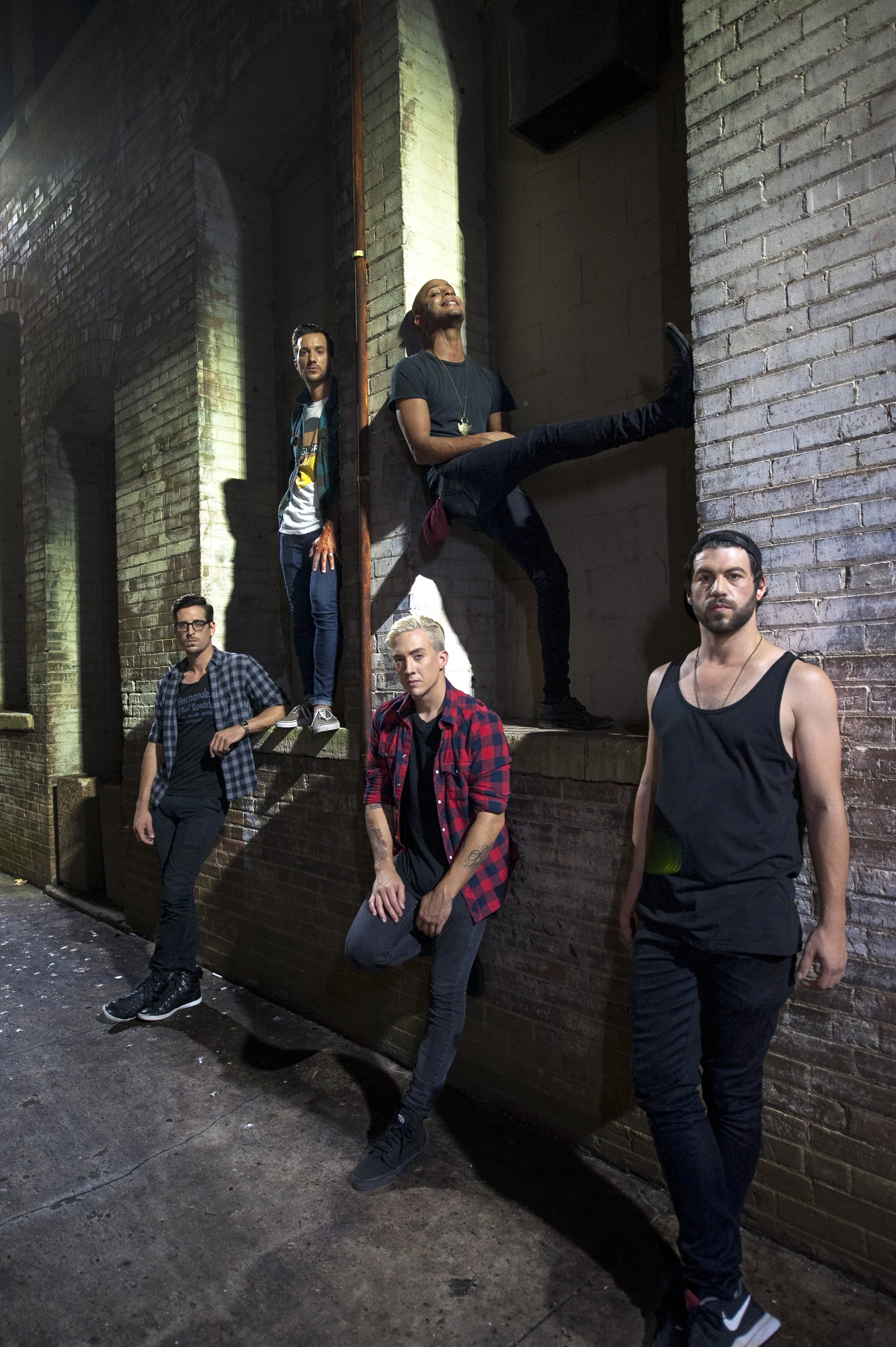 Group of five young adults posing against brick walls in an urban alley, with some standing and some sitting on ledges, at night.