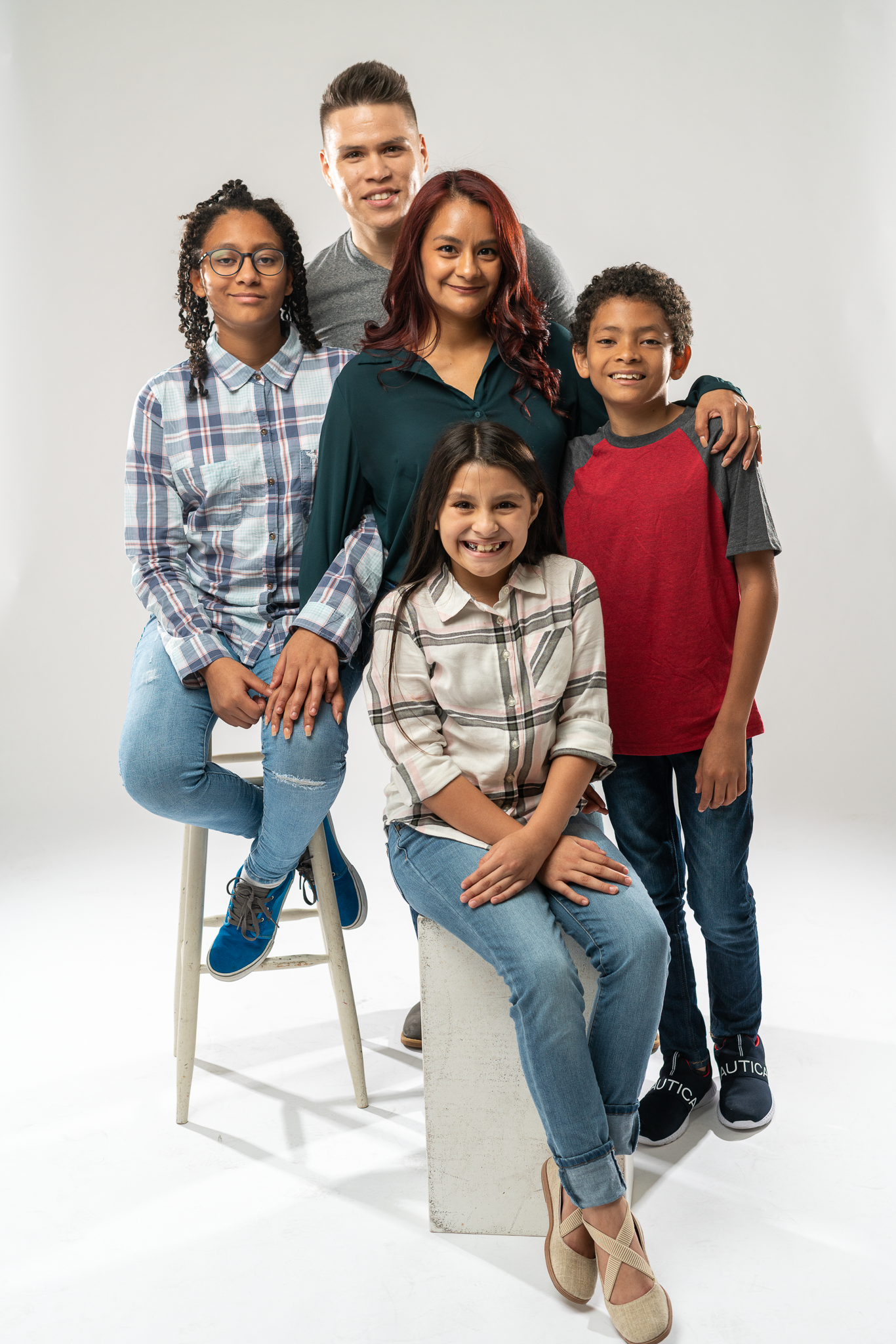 A diverse group of five people, a woman and four children, posing together in a studio against a plain white background. They are smiling and have their arms around each other.