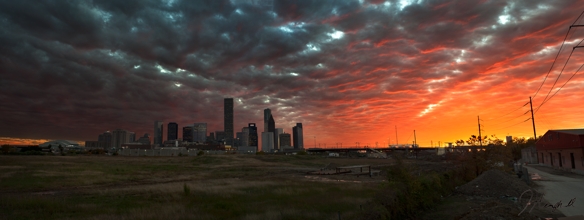 Skyline of a city during sunset with dark clouds overhead and an orange glow near the horizon, with buildings and power lines in the foreground.
