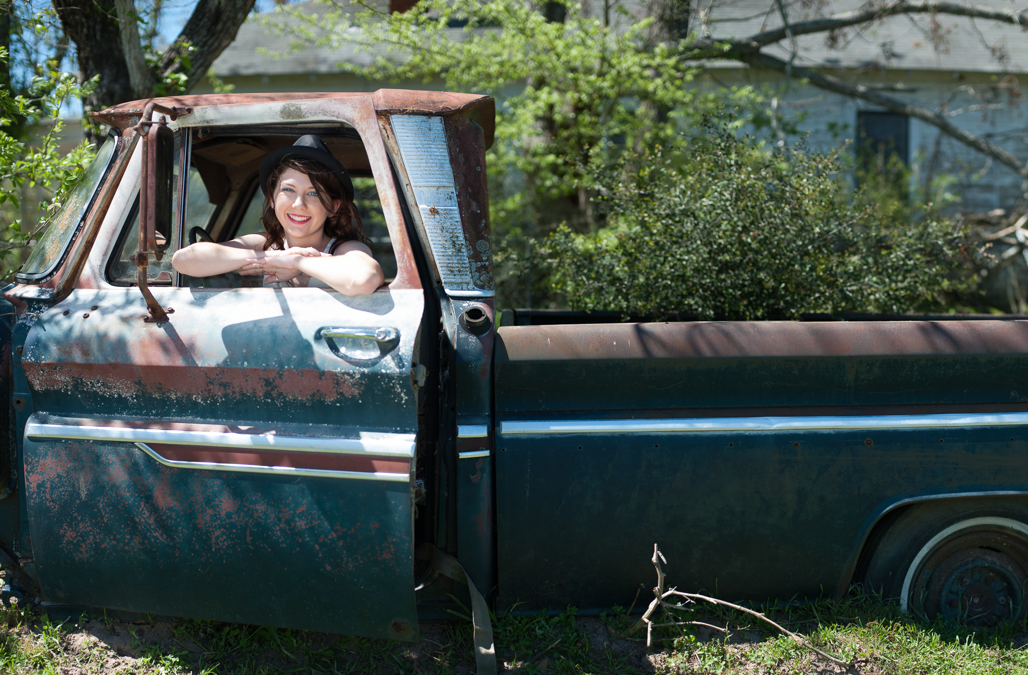 A woman with dark hair, wearing a black hat, smiling, leaning on the open window of an old rusty pickup truck in a yard with green trees and a house in the background.