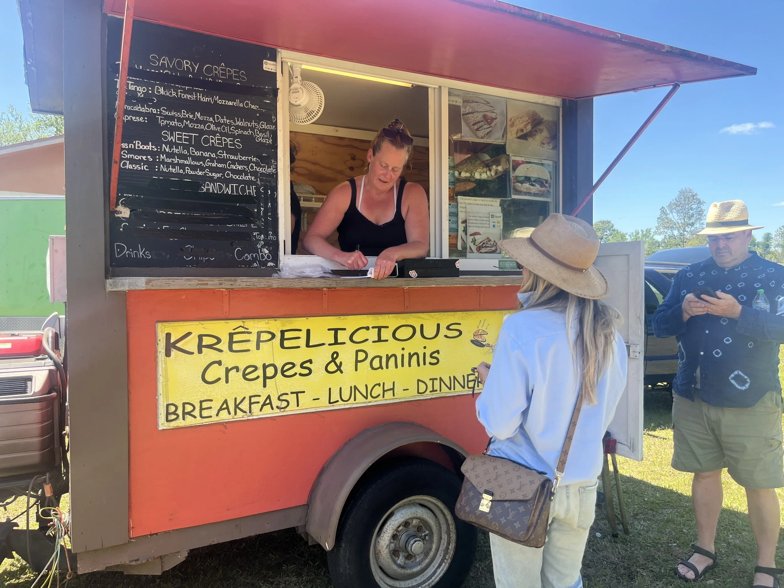 A food truck labeled 'KRÉPELICIOUS Crepes & Paninis' serving breakfast, lunch, and dinner. There is a woman inside the truck taking an order, and two customers standing outside, one woman wearing a wide-brimmed hat and a man in sunglasses and a hat, both using their phones. The truck is outdoors on a sunny day.
