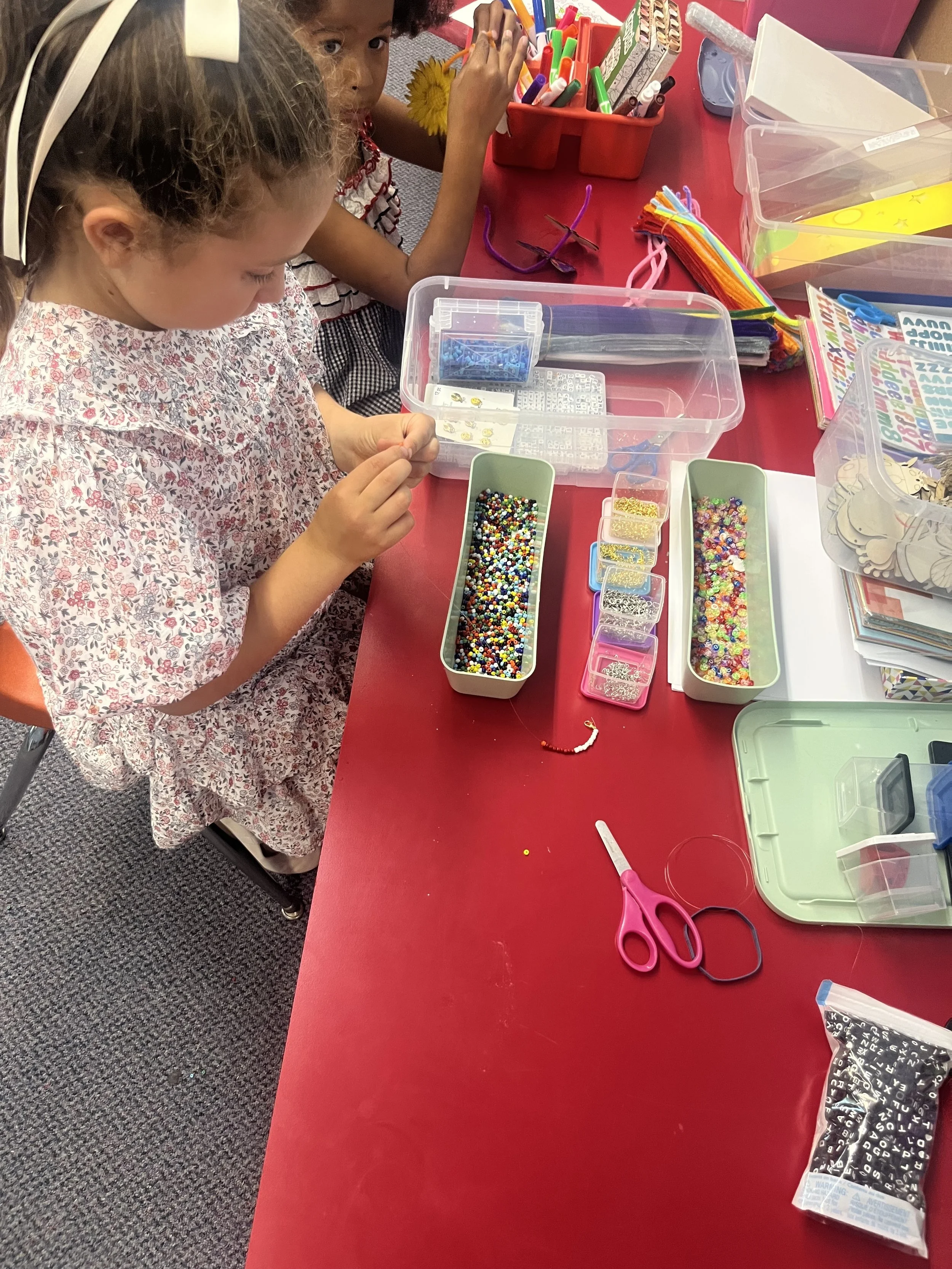 Children sitting at a red table engaged in a craft project with beads, scissors, and containers of craft supplies.