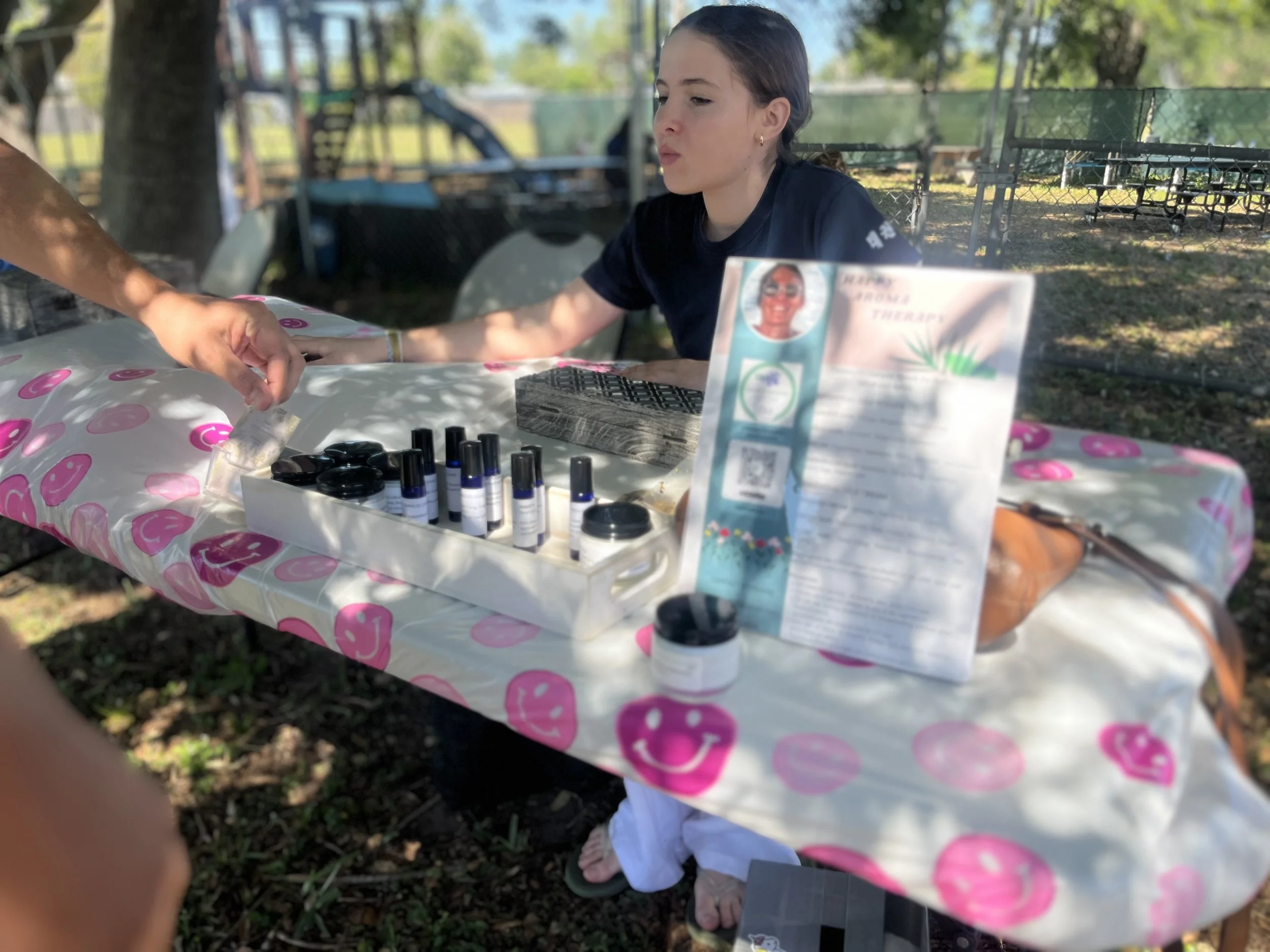 A young woman at an outdoor event sniffs essential oils or perfume samples at a booth with a pink smiley face tablecloth. There is a person handing her a small item, and the booth has bottles, jars, and informational flyers.