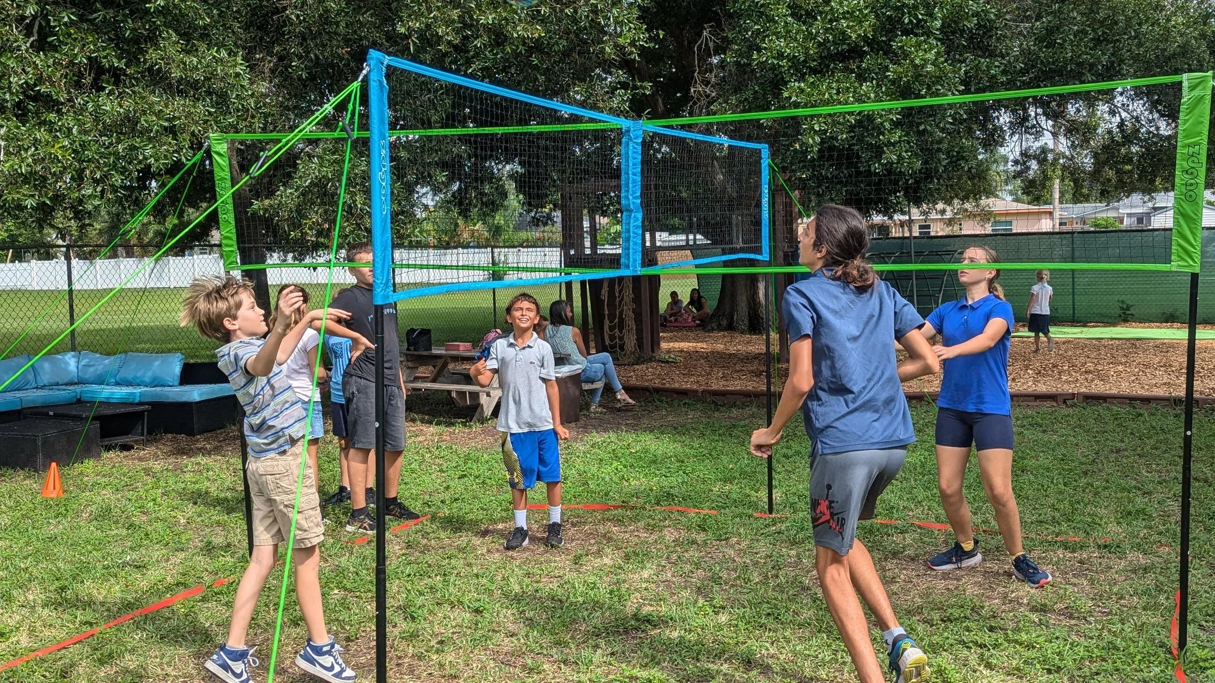 Children playing a game of volleyball in a park, with a volleyball net and green lines marking the court, surrounded by trees and grass.