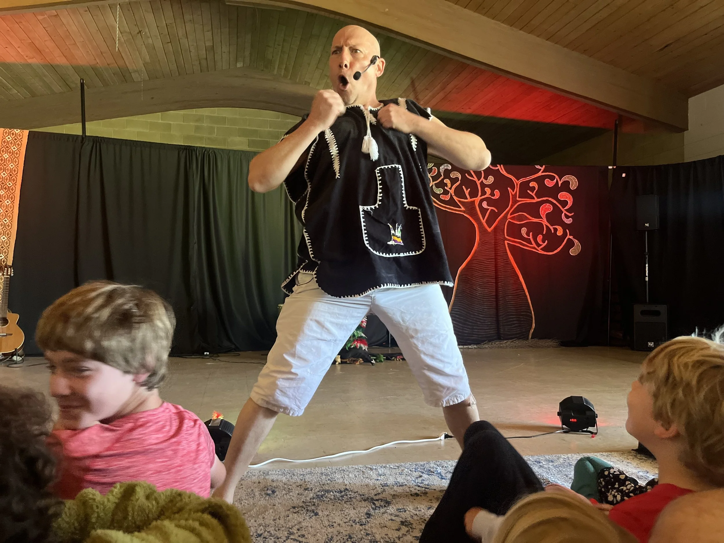 A man performing on stage with a headset microphone, wearing a black shirt with white stitched patterns, beige shorts, and engaging with children seated in front of him. The stage backdrop features a stylized red tree design.