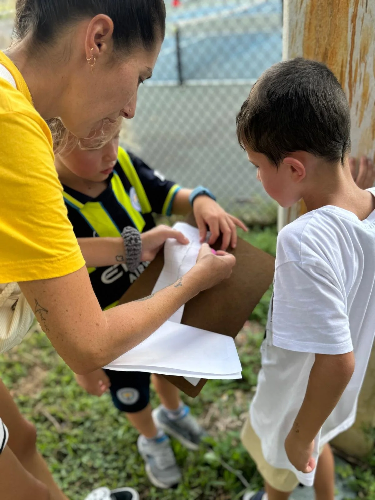A woman with tattoos on her arms signs a piece of paper on a clipboard for a young boy in a white t-shirt, while another child looks on, outdoors near a chain-link fence.