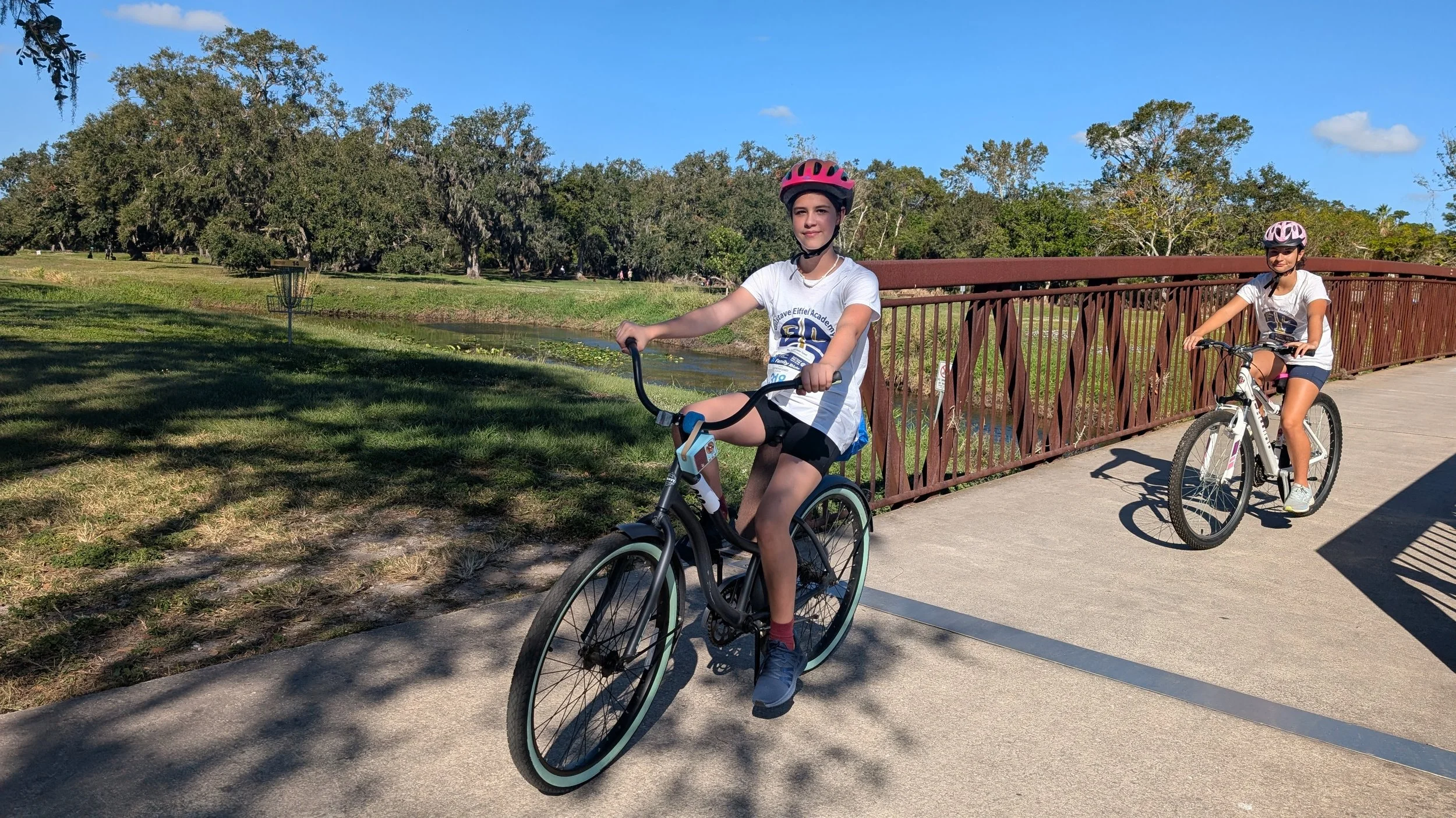 Two young girls riding bicycles on a paved trail in a park, wearing helmets and casual clothes, with trees, a grassy area, a creek, and a brown bridge in the background on a sunny day.