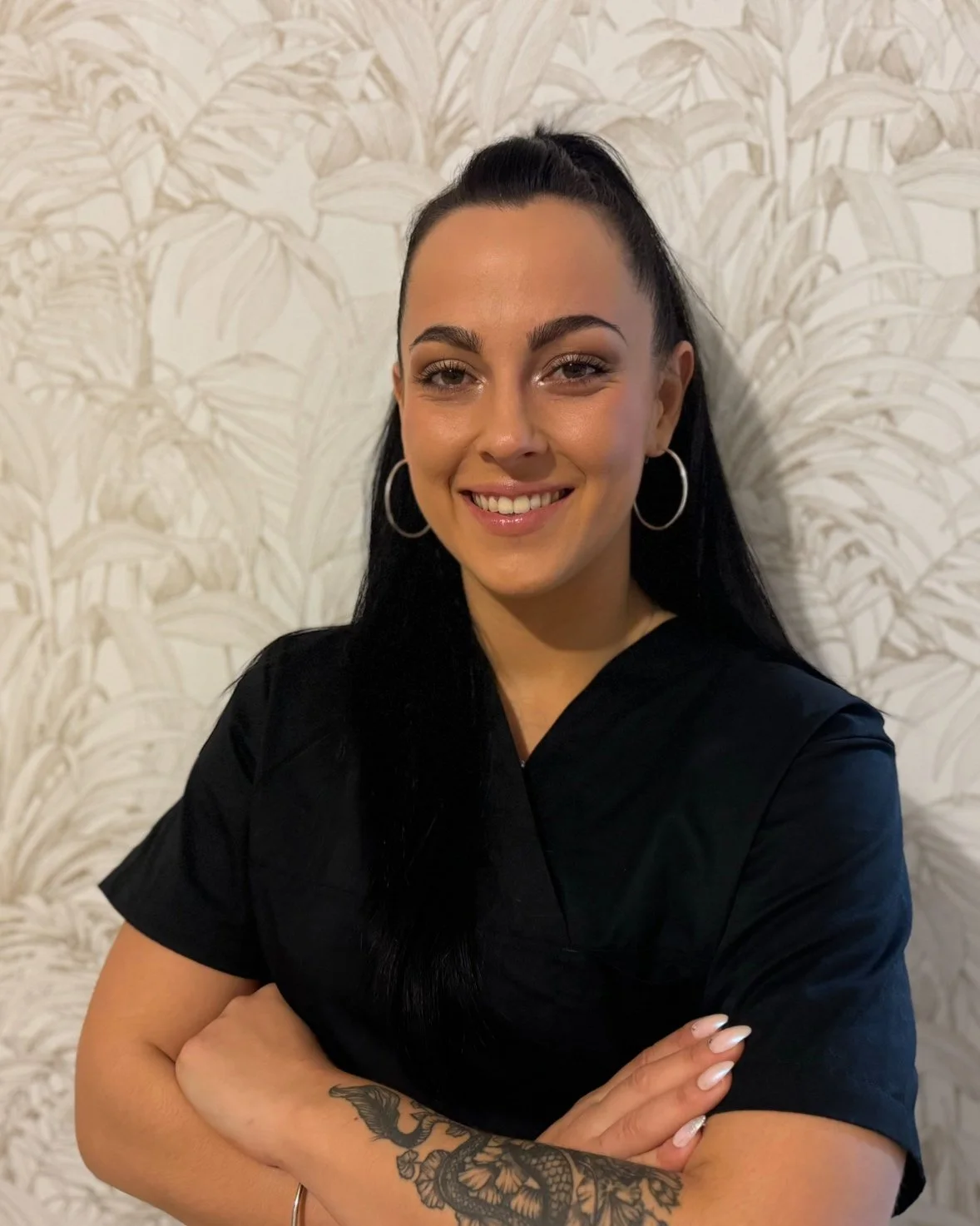 Femme souriante avec boucle d'oreilles en argent, cheveux noirs en queue de cheval, vêtements noirs, tatouage sur l'avant-bras gauche, fond avec motif de feuilles