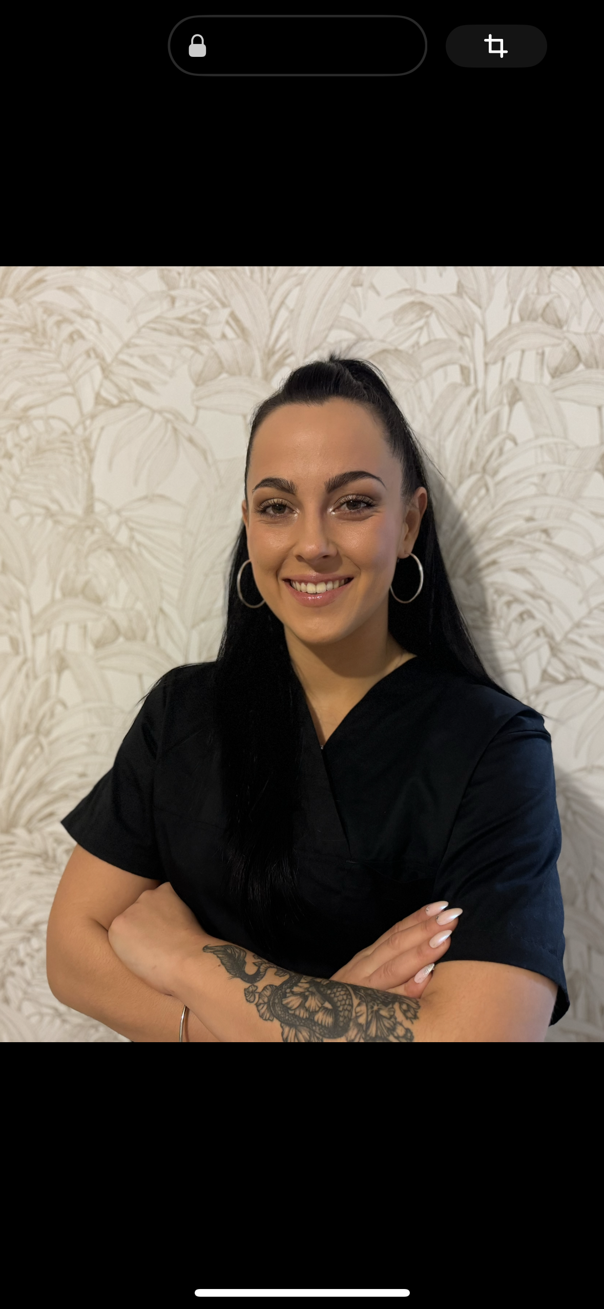 Une femme souriante avec de longs cheveux noirs tirés en arrière, portant de grands anneaux en boucle d'oreille et une robe noire, posant devant un mur à motif feuillage beige.