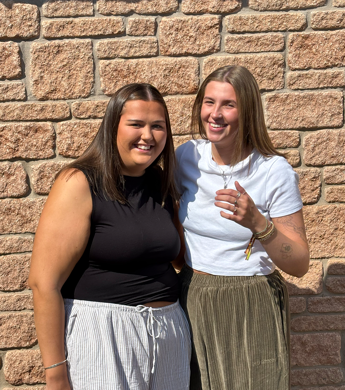 Two young women standing close together in front of a brick wall, smiling at the camera, one with dark hair wearing a sleeveless black top and striped white pants, the other with light hair wearing a white T-shirt and brown striped pants.