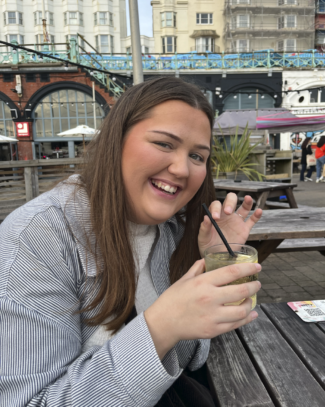 Young woman smiling and holding a beverage with a straw at an outdoor seating area, with a building and string lights in the background.