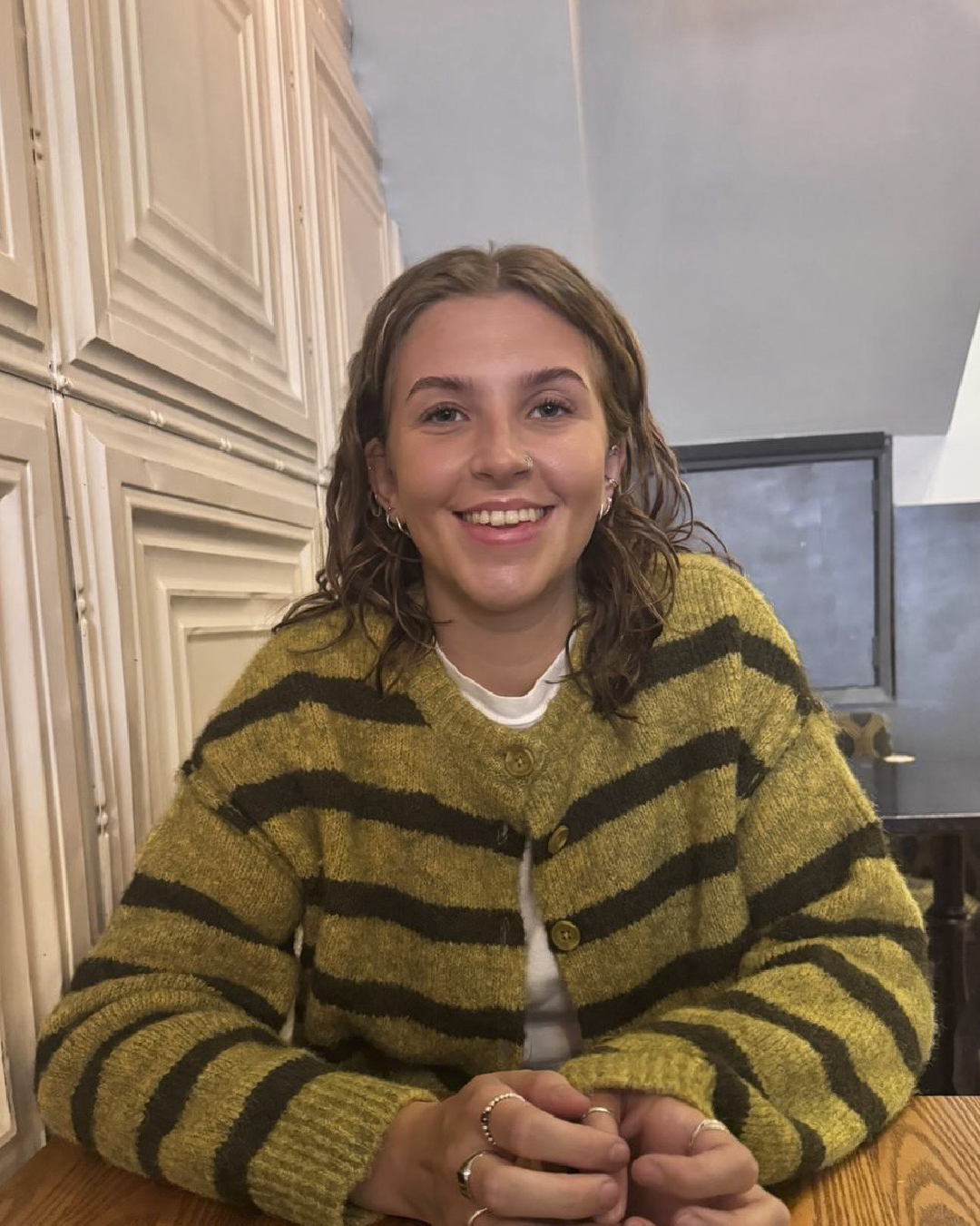 A young woman with curly brown hair and multiple earrings, smiling, sitting at a wooden table in a room with beige cabinets and gray walls, wearing a mustard yellow and black striped sweater.