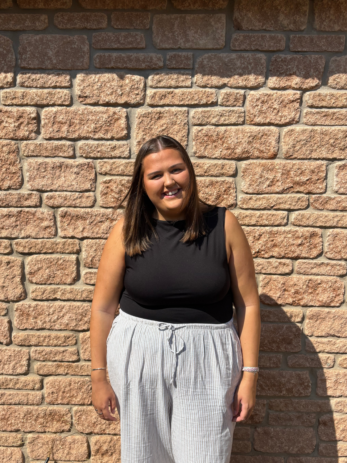 A young woman with shoulder-length brown hair, smiling, standing outdoors against a brick wall. She is wearing a black sleeveless top and light-colored, vertical striped pants with a drawstring.