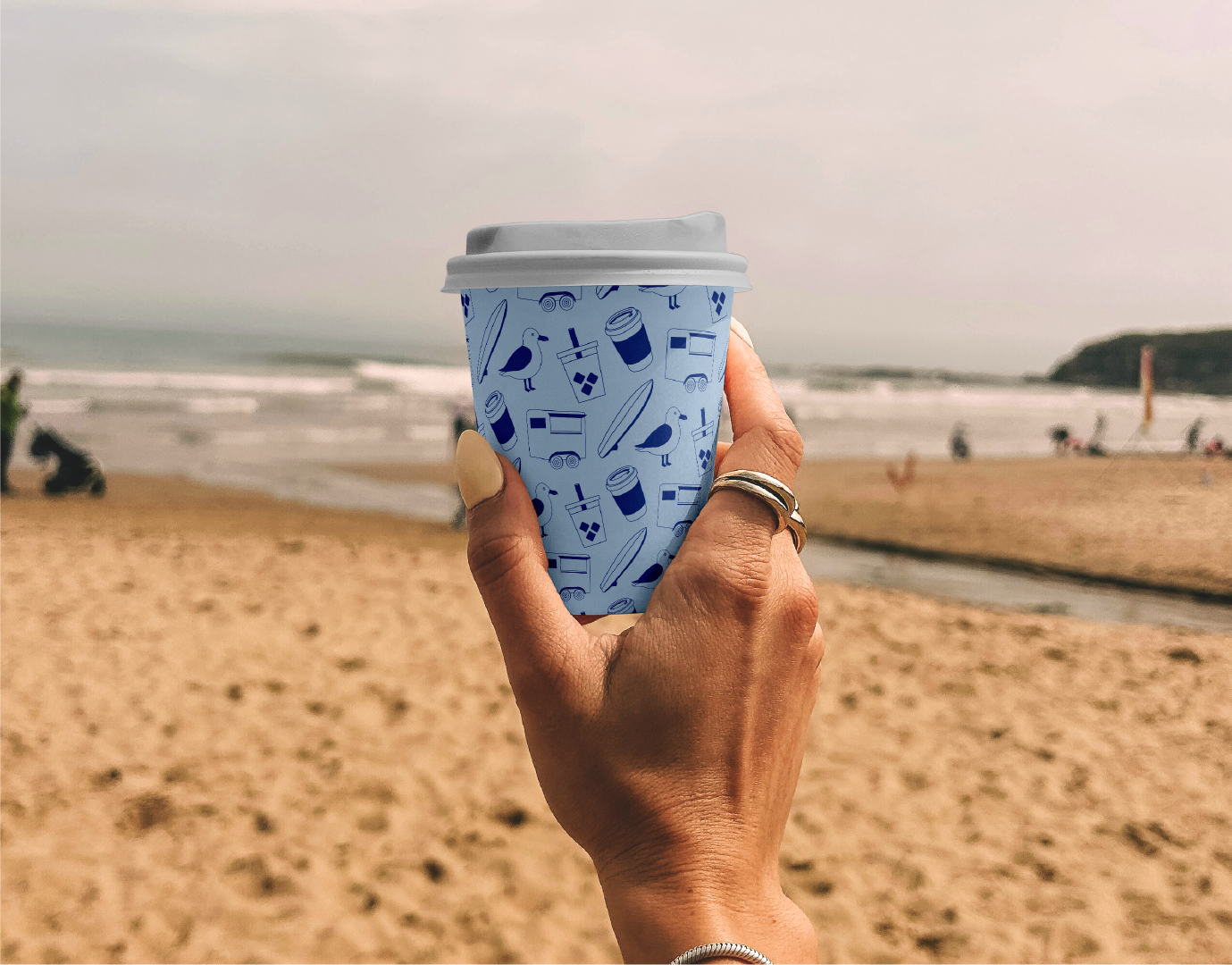 A person holding a disposable coffee cup with a blue pattern featuring seagulls, drink cups, and surfboards on a sandy beach.