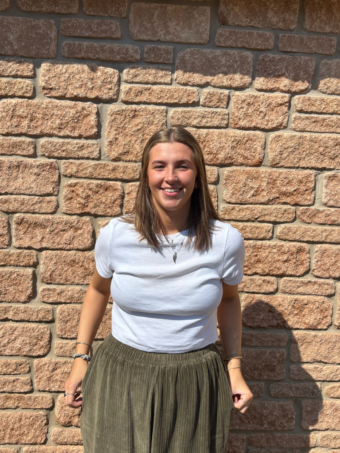A young woman with brown hair and a nose ring smiling, standing against a brick wall, wearing a white t-shirt and olive green pants.
