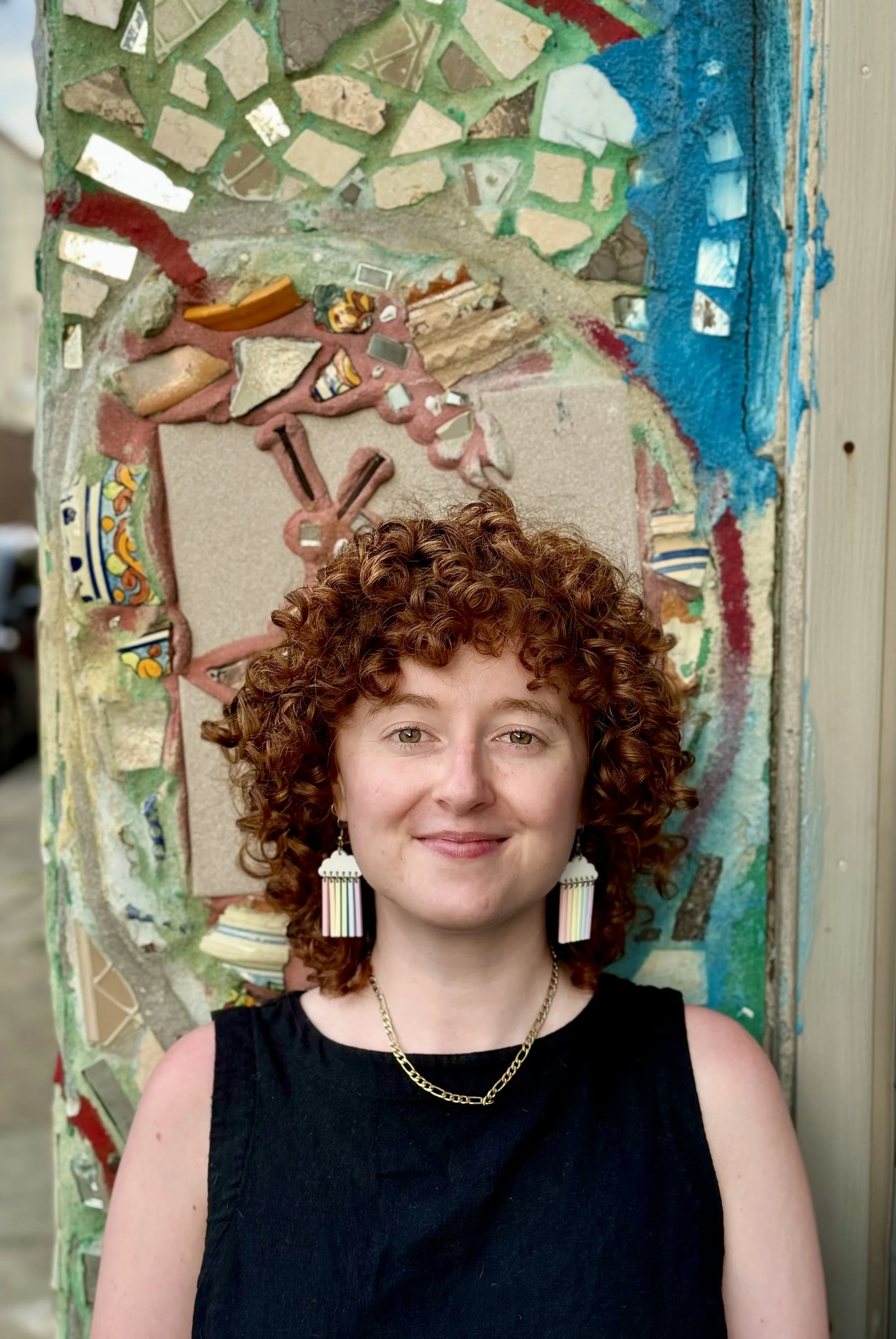 A white non-binary person starts in front of a colorful mosaic mural by Isaiah Zagar. They have curly red hair that is shoulder-length at the sides and cut into bands. They are wearing a gold chain, cloud earrings, and a black sleeveless top.