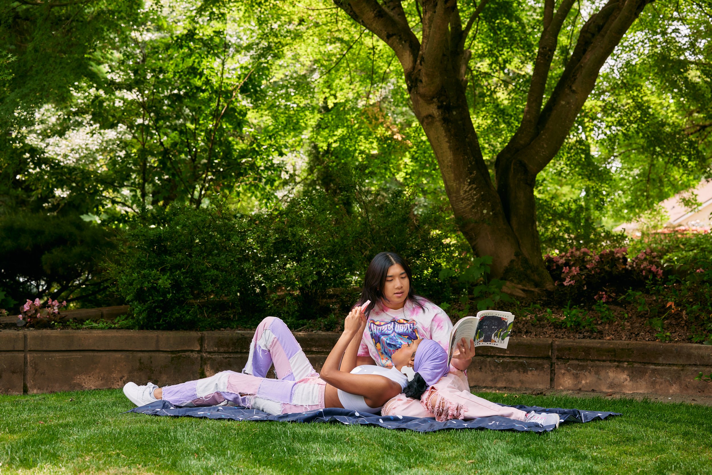 Wide shot of a young autistic couple practicing parallel play while lounging on a picnic blanket at a lush green park. On the left, the Black woman lies on her partner’s lap, concentrating on her fidget toy. The non-binary Asian person reads a book.