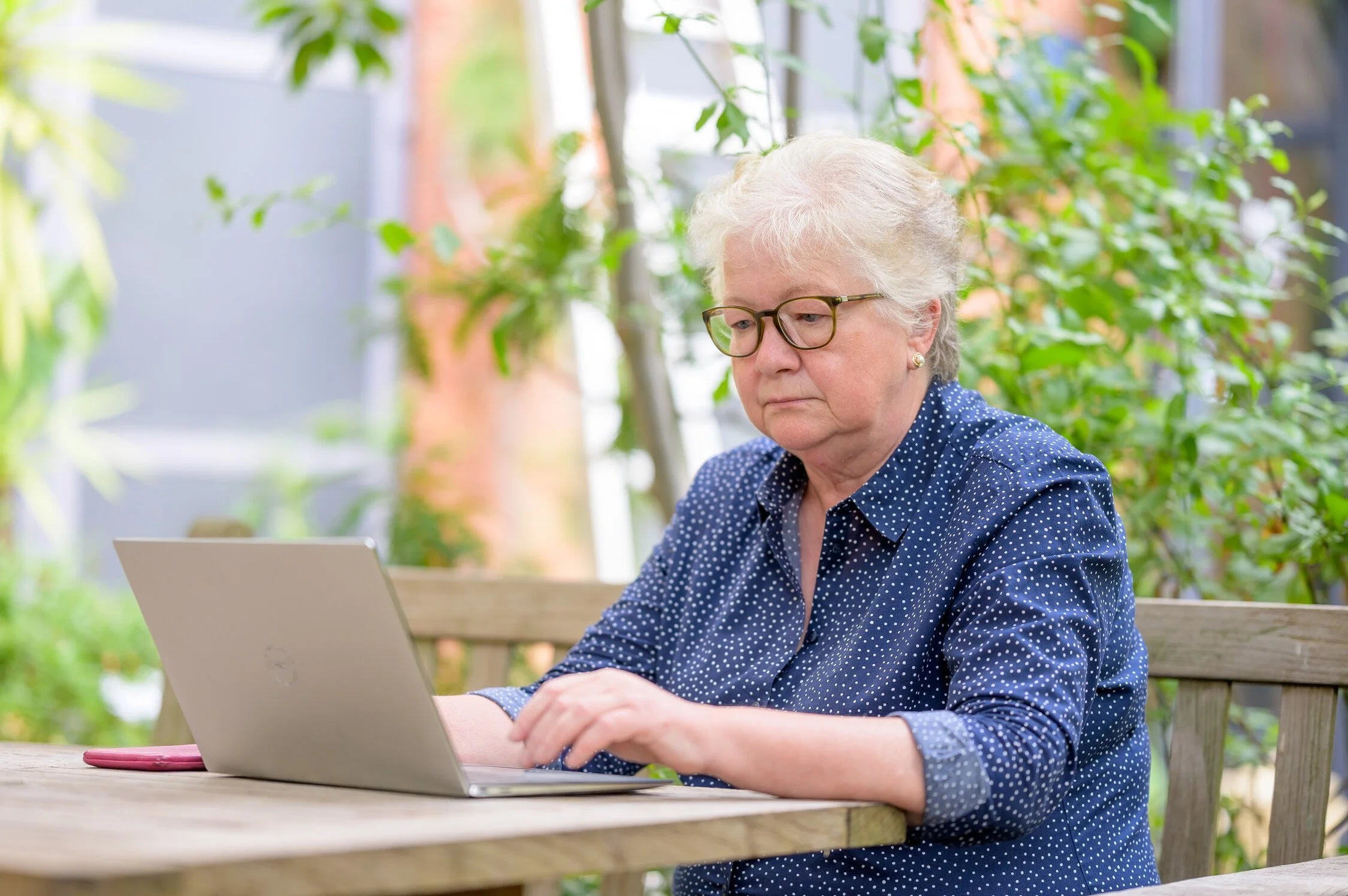 A white woman over fifty looks at a silver laptop. The woman is white with white hair and is wearing a blue collared shirt with small white dots. She is wearing glasses. Image provided by agewithoutlimits.org.