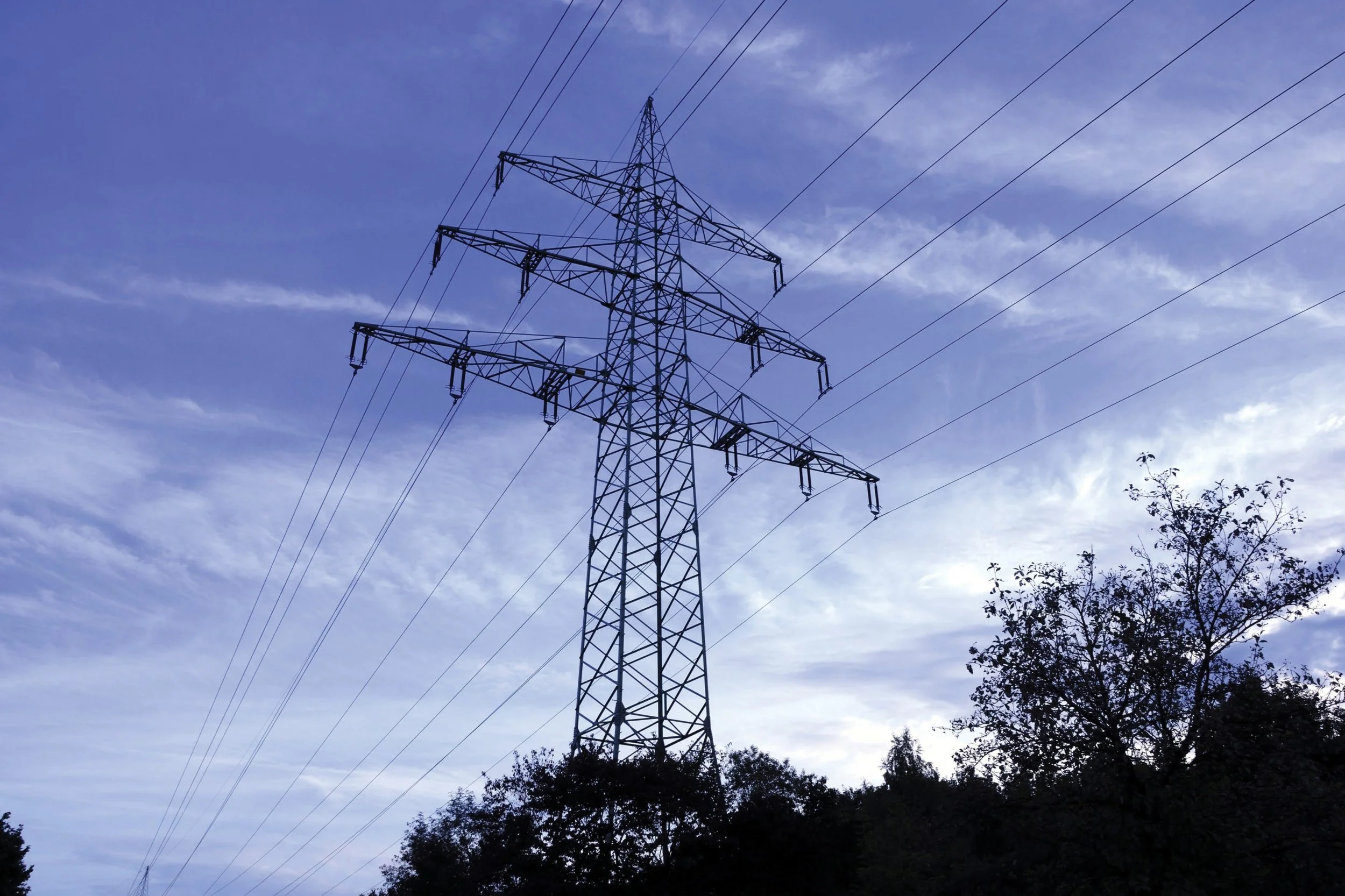 A large electrical utility pole with wires against a blue sky with clouds and trees