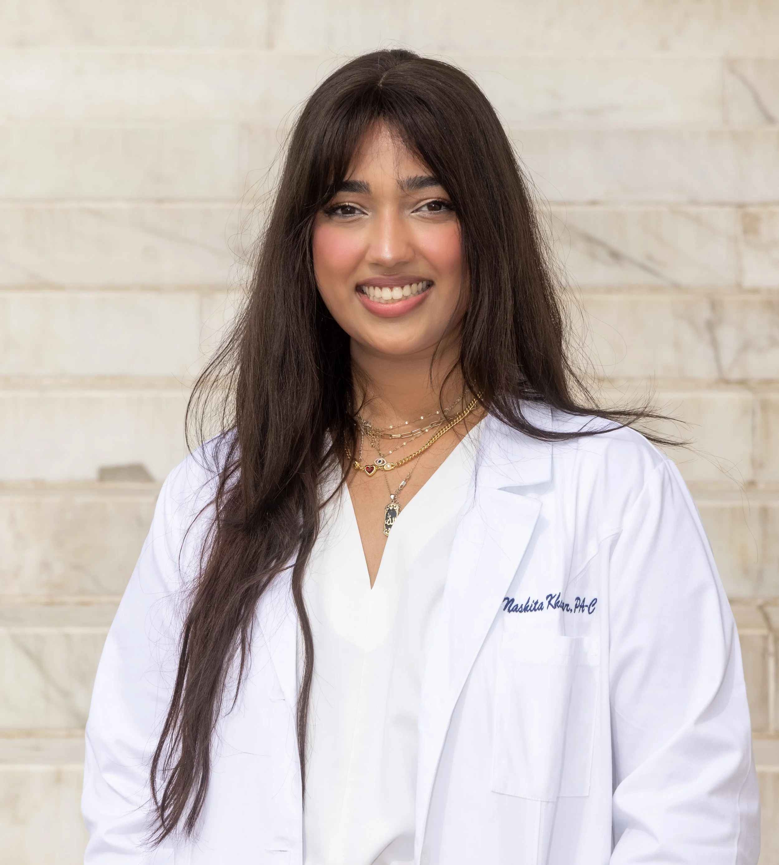 A woman in a white medical coat smiling, standing in front of a light beige brick wall.
