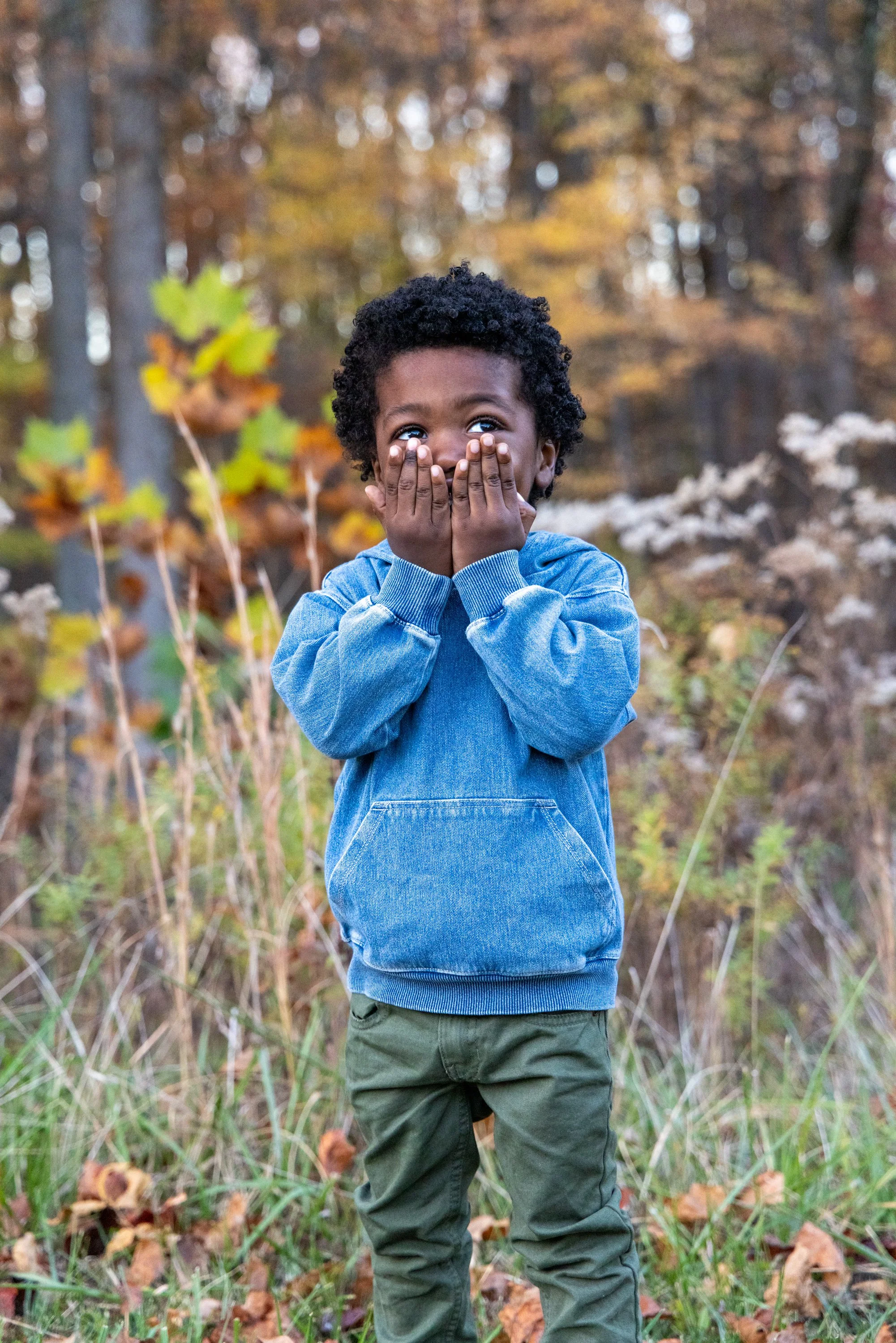 A young boy with curly black hair covering his mouth with his hands, standing outdoors in a wooded area with autumn leaves, wearing a blue hoodie and green pants.