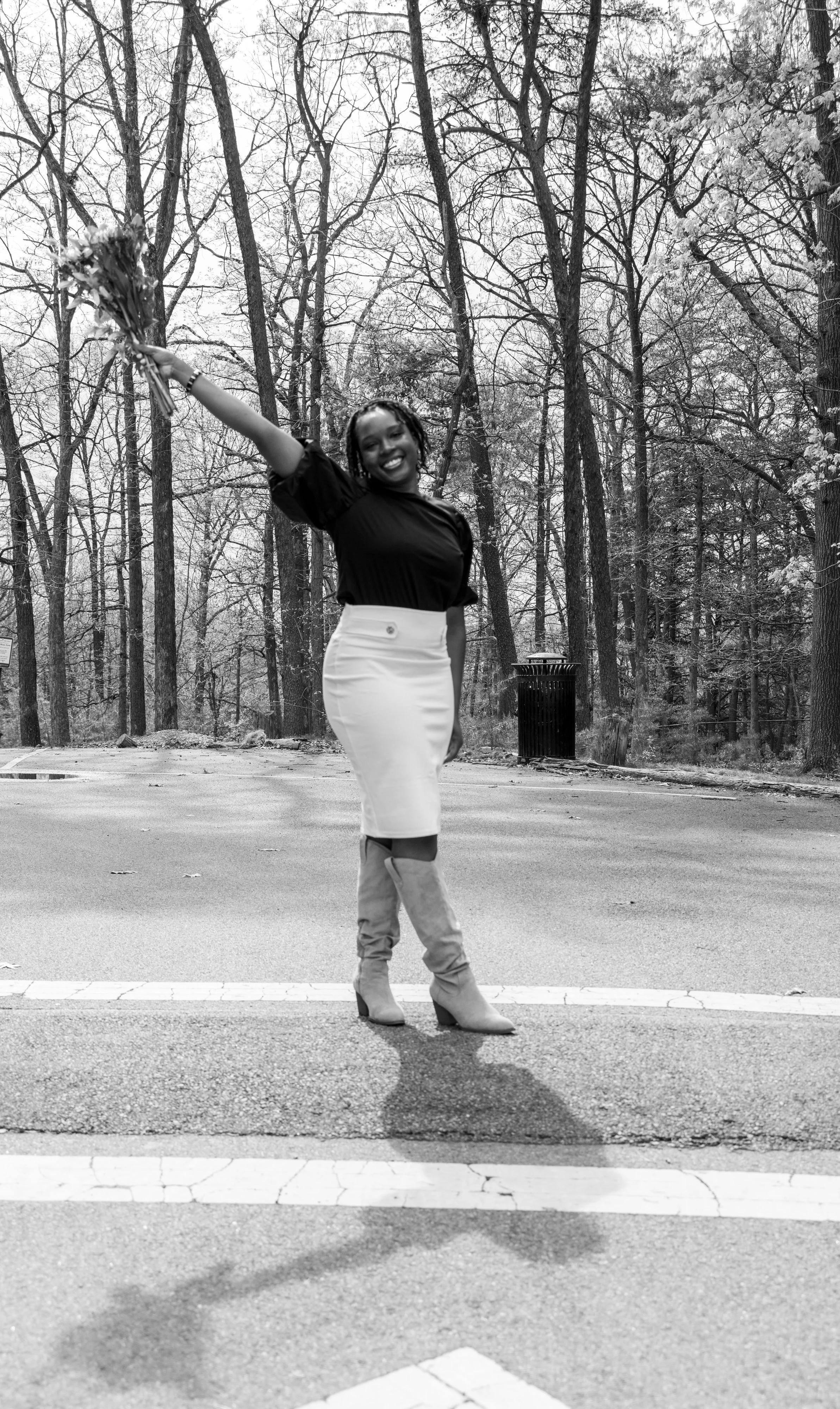 A woman standing on a crosswalk in a park with bare trees, holding a bouquet of flowers raised in the air and smiling.