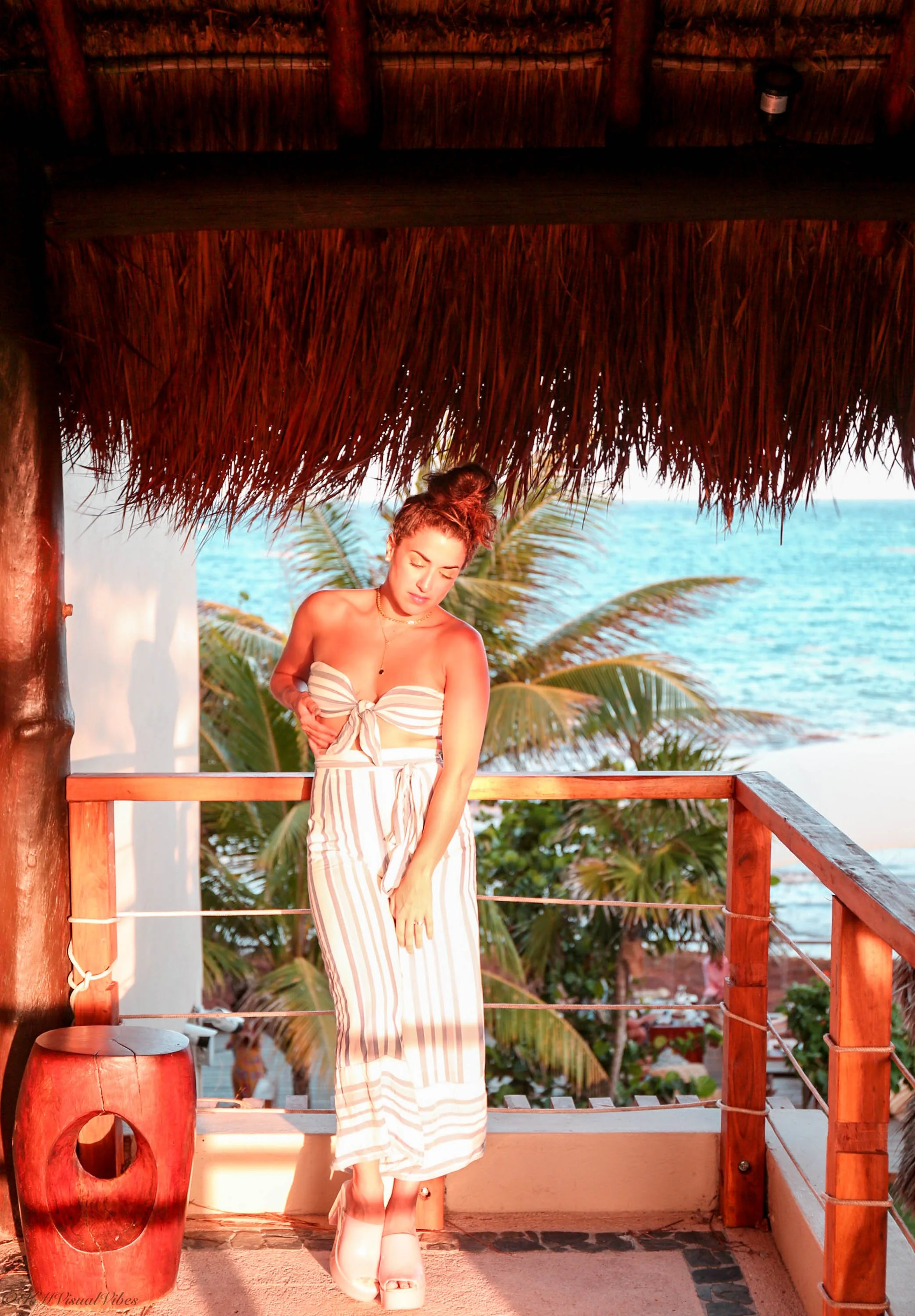 A woman in a striped summer dress and platform shoes standing on a balcony with a thatched roof, overlooking palm trees, ocean, and sky at sunset.