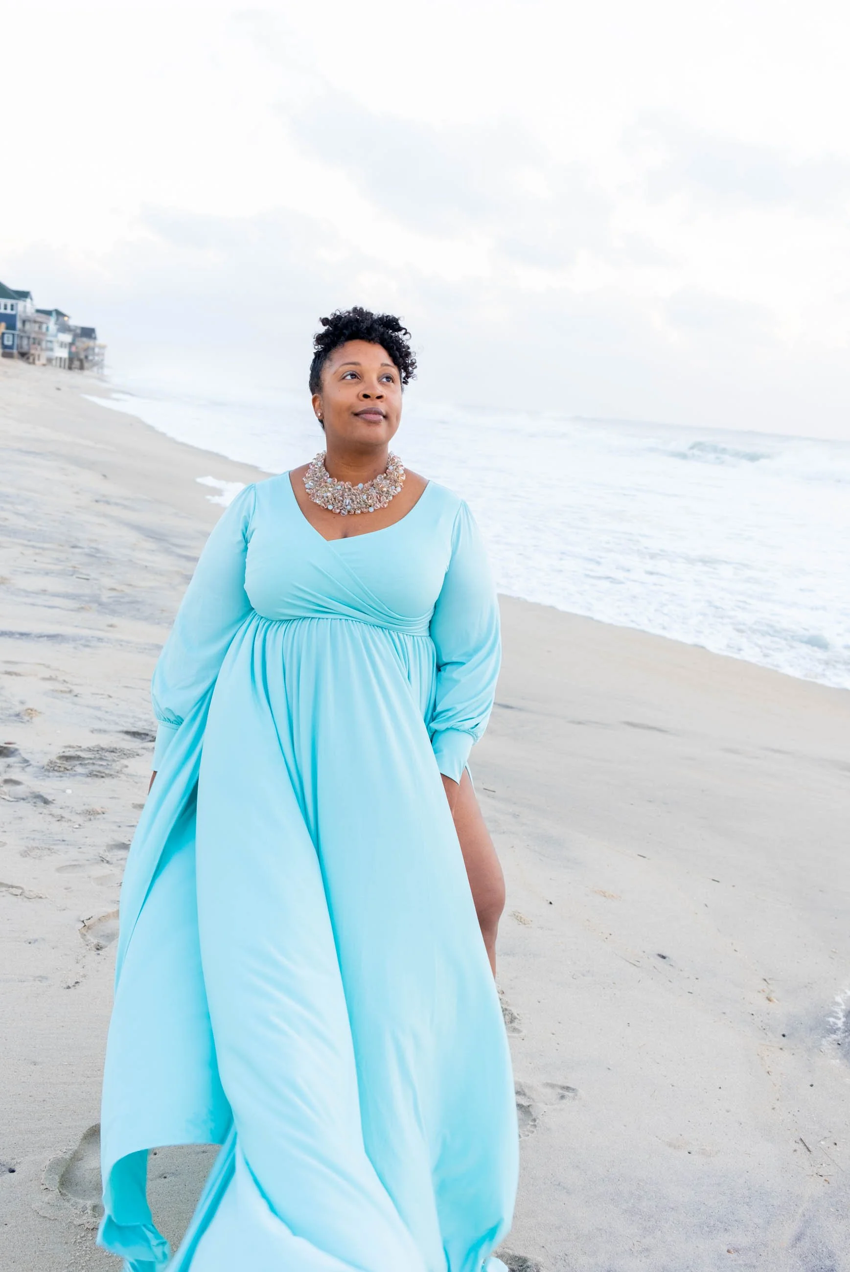 A woman in a flowing light blue dress with a statement necklace walking along the sandy beach with ocean waves in the background.