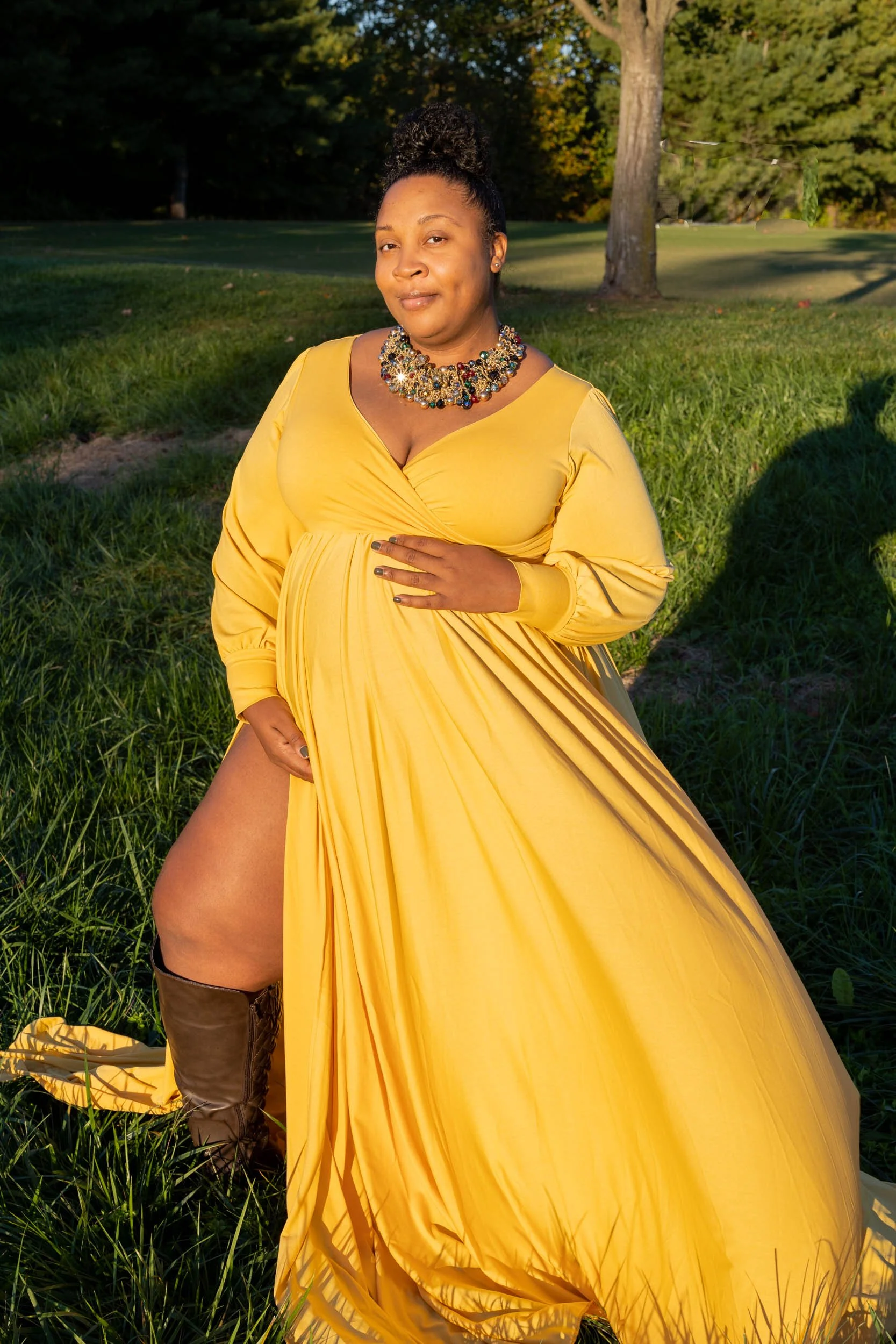 A woman in a long yellow gown with a high slit, wearing a statement necklace, standing outdoors in a grassy area with trees in the background.